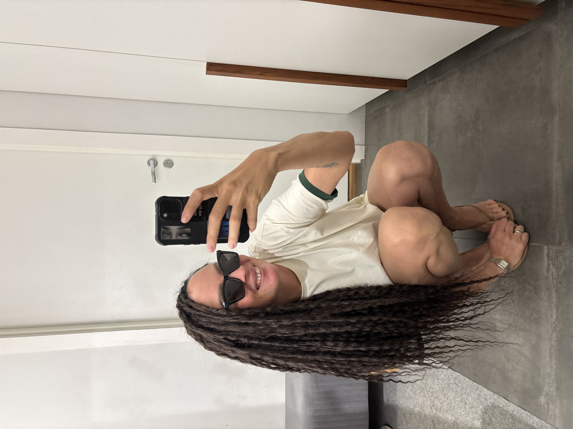 An individual with long, dark brown dreadlocks and black square-framed sunglasses is depicted in an indoor setting in Chaikovsky, Russia. The person, who appears to be female, is crouching or squatting on a dark gray tiled or polished concrete floor, holding a black smartphone in their right hand to take a mirror selfie. They are smiling, revealing their teeth. The individual wears a light cream-colored short-sleeved t-shirt with a dark green trim on the sleeves, light-colored shorts partially visible, and light brown flip-flops. A silver-toned watch with a metal band is worn on the left wrist, along with a silver-toned ring on the fourth finger of the left hand. The background features white walls, with a white door on the left equipped with a silver-toned handle and lock. A low, gray upholstered bench or furniture piece is visible on the far left. On the right side, a vertical wooden trim of medium brown frames a white panel or surface. The environment appears clean, modern, and well-lit.