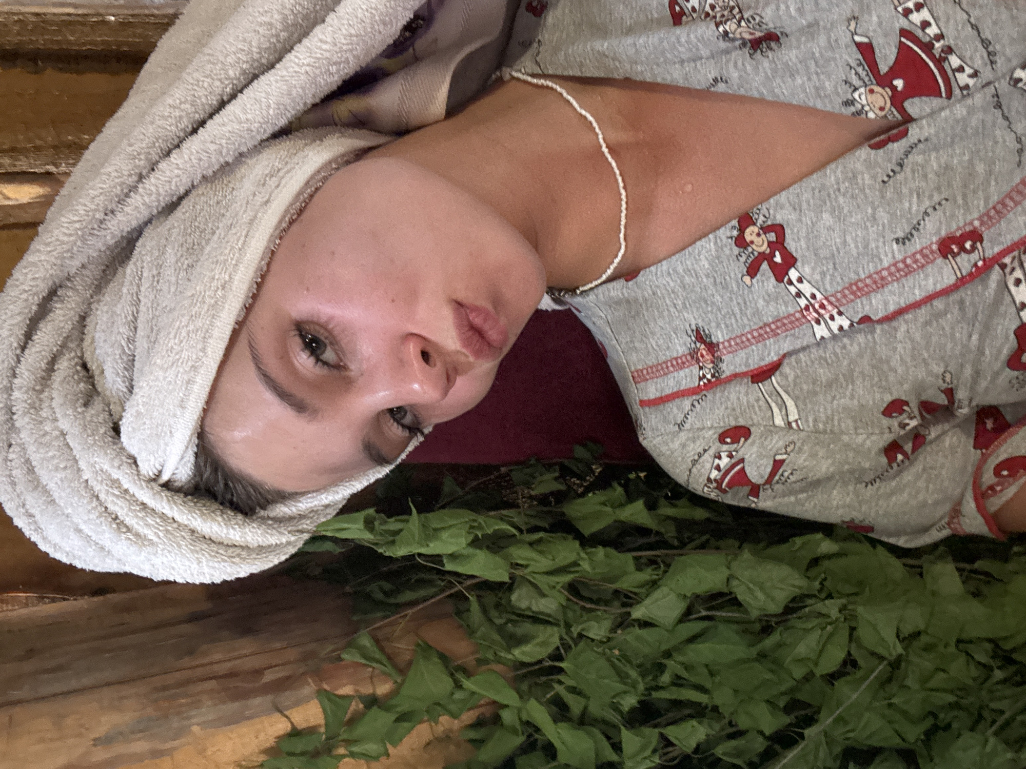 A woman with a white towel wrapped around her head is lying in what appears to be a sauna in Chaikovsky, Russia. She is wearing a grey pajama top with a red and white pattern. There are green leaves and branches on the floor next to her. She is wearing a white beaded necklace. The sauna walls are made of wood.