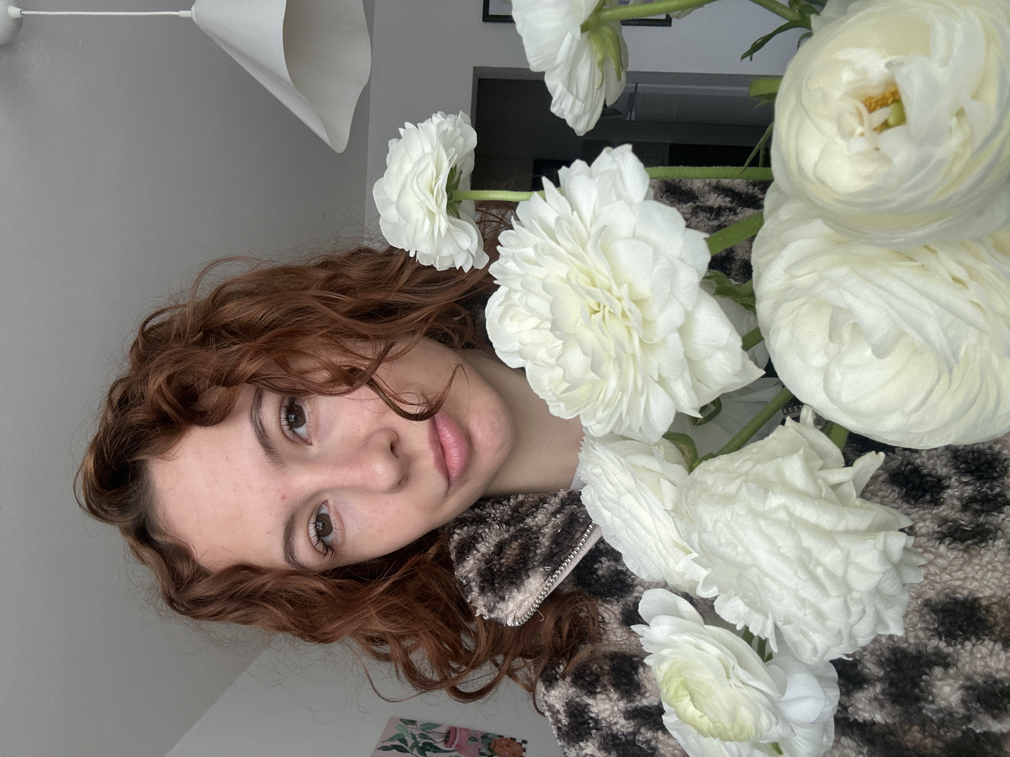A young woman with reddish-brown curly hair and dark eyes is depicted in a close-up portrait, rotated 90 degrees counter-clockwise. Her head and upper torso are visible on the left side of the frame, with her gaze directed towards the viewer, conveying a calm and gentle expression. She has fair skin and naturally pink lips. She wears a jacket with a distinct brown, black, and cream animal print pattern, featuring a visible silver zipper pull.
A prominent bouquet of white ranunculus flowers dominates the right side of the image, partially obscuring the woman's hair and clothing. The flowers showcase densely packed, ruffled white petals and visible green stems. One flower near the top right reveals a glimpse of its yellowish-green center.
The setting is indoors, likely a home in Novi Sad, Serbia, with a plain light gray wall serving as the main background. In the upper left, a white, sculptural light fixture hangs from the ceiling. A small portion of a framed artwork with light green and pink elements is visible in the lower left background, beneath the woman's chin. A dark, rectangular object can be subtly seen in the upper right background, behind some of the flowers. The lighting is soft and even, indicating daytime conditions indoors. The woman is posing directly for the camera. No discernible text is present in the image.