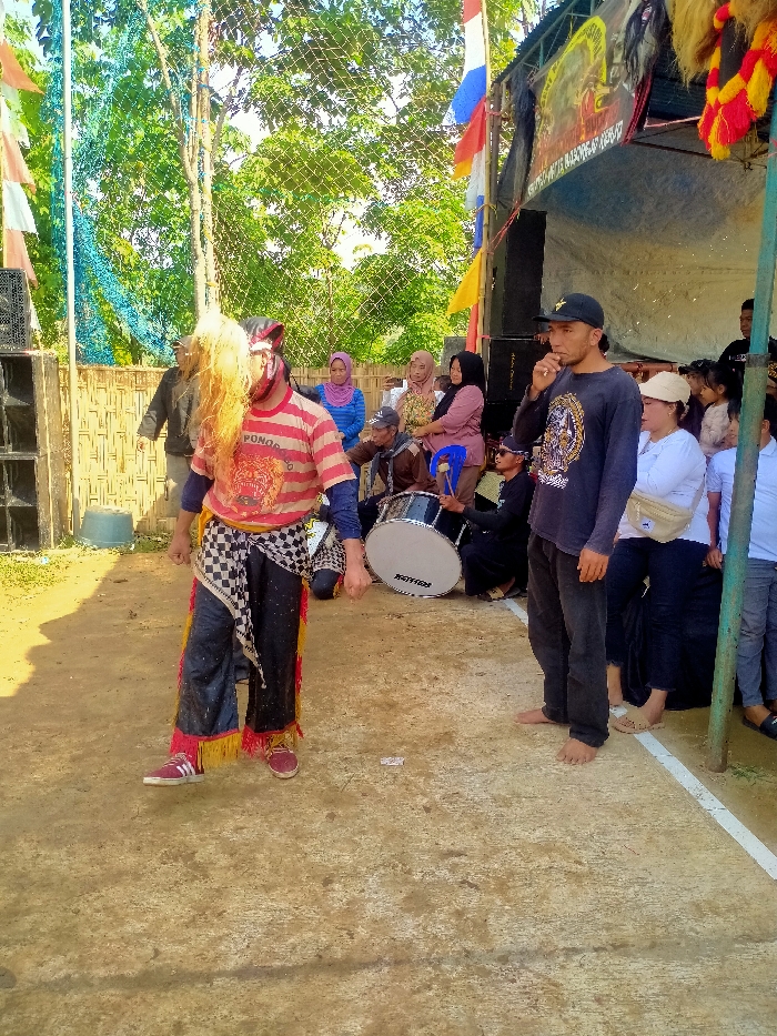 The image depicts an outdoor event in Plosorejo, Indonesia, under natural daylight. A male individual, positioned centrally, is attired in performance wear: a red and white striped t-shirt featuring a tiger graphic and the text "PONOROG...", dark long-sleeved undershirt, dark pants with red and yellow fringed ankles, and a black and white checkered fabric tied around the waist. His face is obscured by a mask or heavy makeup, and he wears a long, light-colored wig. He is wearing red and white athletic shoes and has his left foot slightly elevated.
To the right of the central figure, a barefoot male stands facing left, wearing a dark long-sleeved shirt with a graphic and a black baseball cap. His right hand is raised towards his mouth.
Behind these two figures, a group of approximately ten individuals is situated, some seated and some standing. This group includes multiple women, several wearing headscarves, and men. One man is seated near a white 'MOTION' brand bass drum and holds a drumstick.
The background features a makeshift performance area with a light-colored canopy, from which banners with text and graphics are hung. A large dark speaker is visible on the left, adjacent to a temporary fence constructed from bamboo and mesh. Red, white, blue, and yellow flags or streamers are visible. Beyond the immediate performance area, lush green trees and foliage are present. The ground consists of light brown earth and a concrete section marked by a distinct white horizontal line. The scene is brightly lit, casting visible shadows.