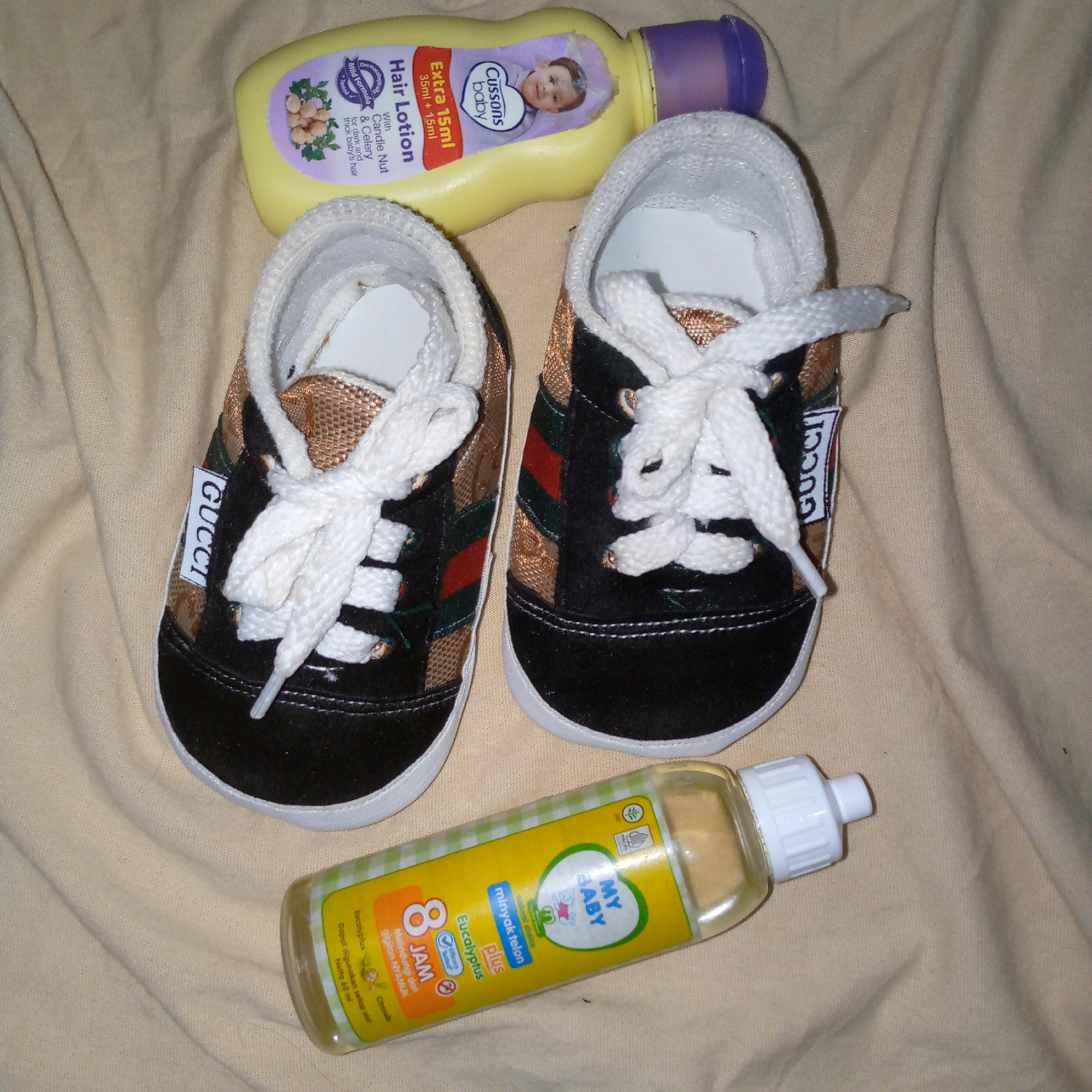 A flat lay photograph displays a collection of baby-related items on a wrinkled, light beige fabric surface. In the upper part of the frame, a yellow bottle of "Cussons baby Hair Lotion" with a purple screw cap is oriented horizontally, angled slightly towards the upper right. The label indicates "Extra 15ml 35ml + 15ml" and features text specifying "Dermatologically Mild Formula Tested" and "With Candle Nut & Celery for dark and thick baby's hair," alongside an image of a baby's face. Centered below the lotion is a pair of black and brown baby shoes, positioned side-by-side with their toes pointing towards the top of the image. The shoes feature white rubber soles and white laces, which are tied. The main body of the shoes is black with a patterned brown/gold fabric section on the sides, accented by distinct red and green stripes. A white tag, vertically displaying "GUCCI" in black text, is attached to the outer side of the left shoe. In the lower portion of the image, a clear plastic bottle of "MY BABY minyak telon plus" with a white screw cap is placed horizontally, angled slightly towards the upper left. Its yellow label includes a graphic of a baby, text stating "8 JAM" (8 hours) in red, and mentions "Eucalyptus" and "Citronella," with additional text in Indonesian indicating "Melindungi dari gigitan NYAMUK" (Protects from mosquito bites) and "Netto 60 ml." No individuals or actions are present. The visual content does not provide information to confirm the location as Meunasahblang, Indonesia.