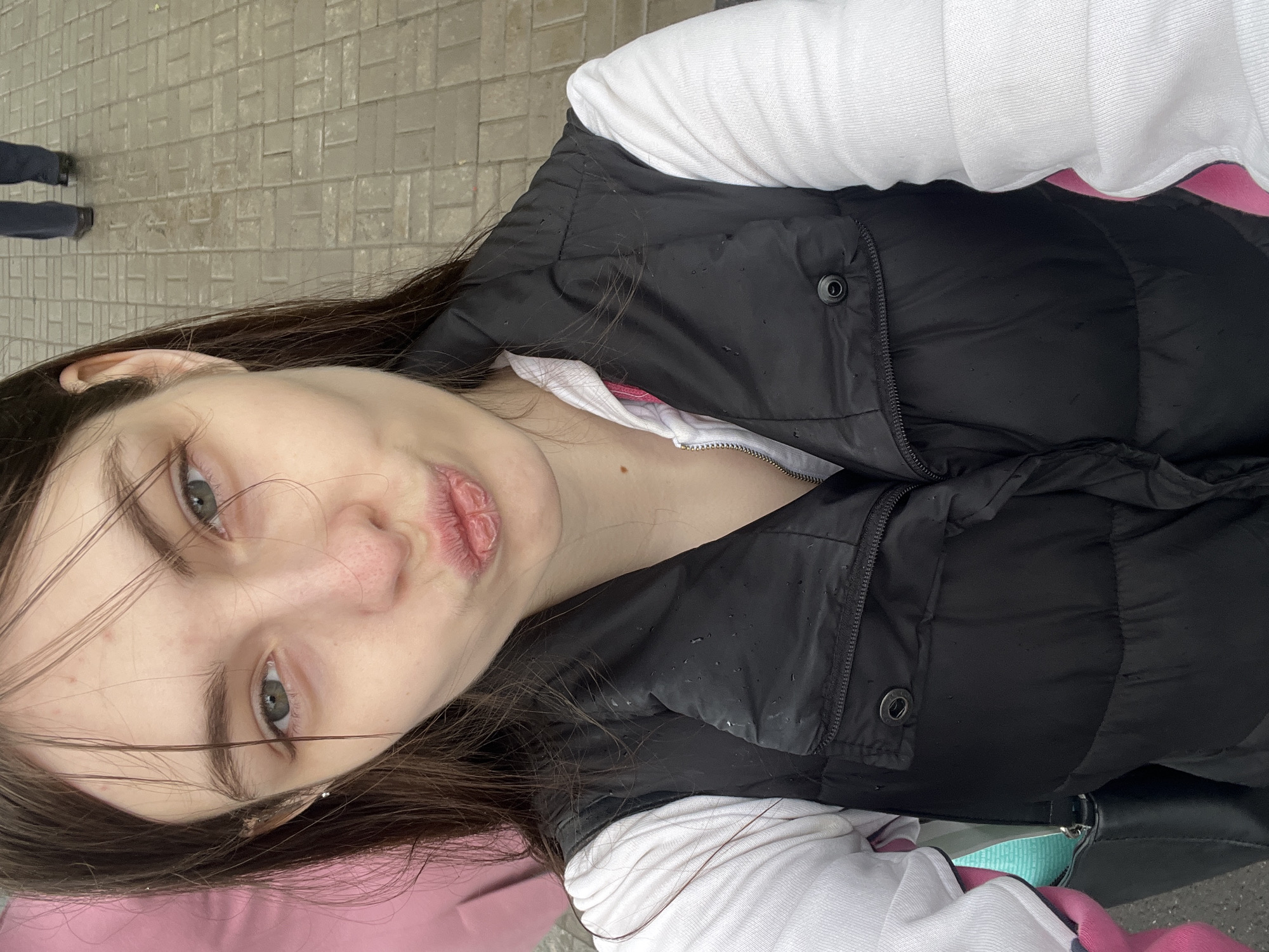 A young woman is taking a selfie on a brick-paved surface. She has brown hair and is wearing a black puffer vest over a white long-sleeved shirt with a hint of pink underneath. Her lips are puckered as if she is about to kiss. In the background, a portion of another person's legs can be seen. There is no information to conclude she is in Saint Petersburg.