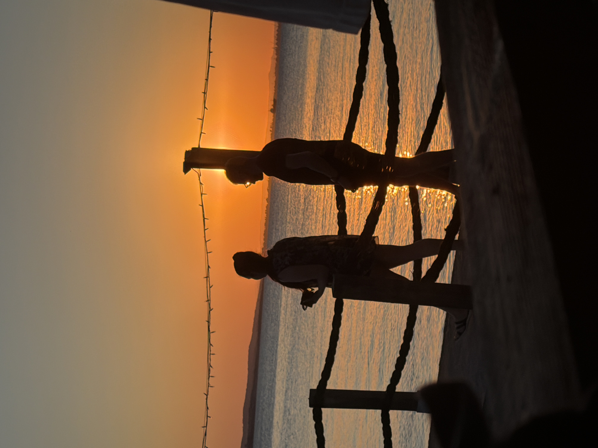 A silhouetted couple walks along a sandy path beside a body of water, likely the Black Sea, as the sun sets over the horizon, casting a warm orange glow across the sky and water. The couple is positioned near a rustic wooden fence made of posts and thick rope, which separates the path from the water. A string of small lights is strung above the fence, illuminated against the darkening sky. The man on the right is wearing glasses and a t-shirt, while the woman on the left is wearing a patterned top and appears to be holding something in her hand. The background shows a distant shoreline with faint hills. The scene conveys a sense of peace and romanticism associated with a sunset by the sea, possibly at the Burgas Sea Garden in Burgas, Bulgaria, known for its scenic waterfront promenade.