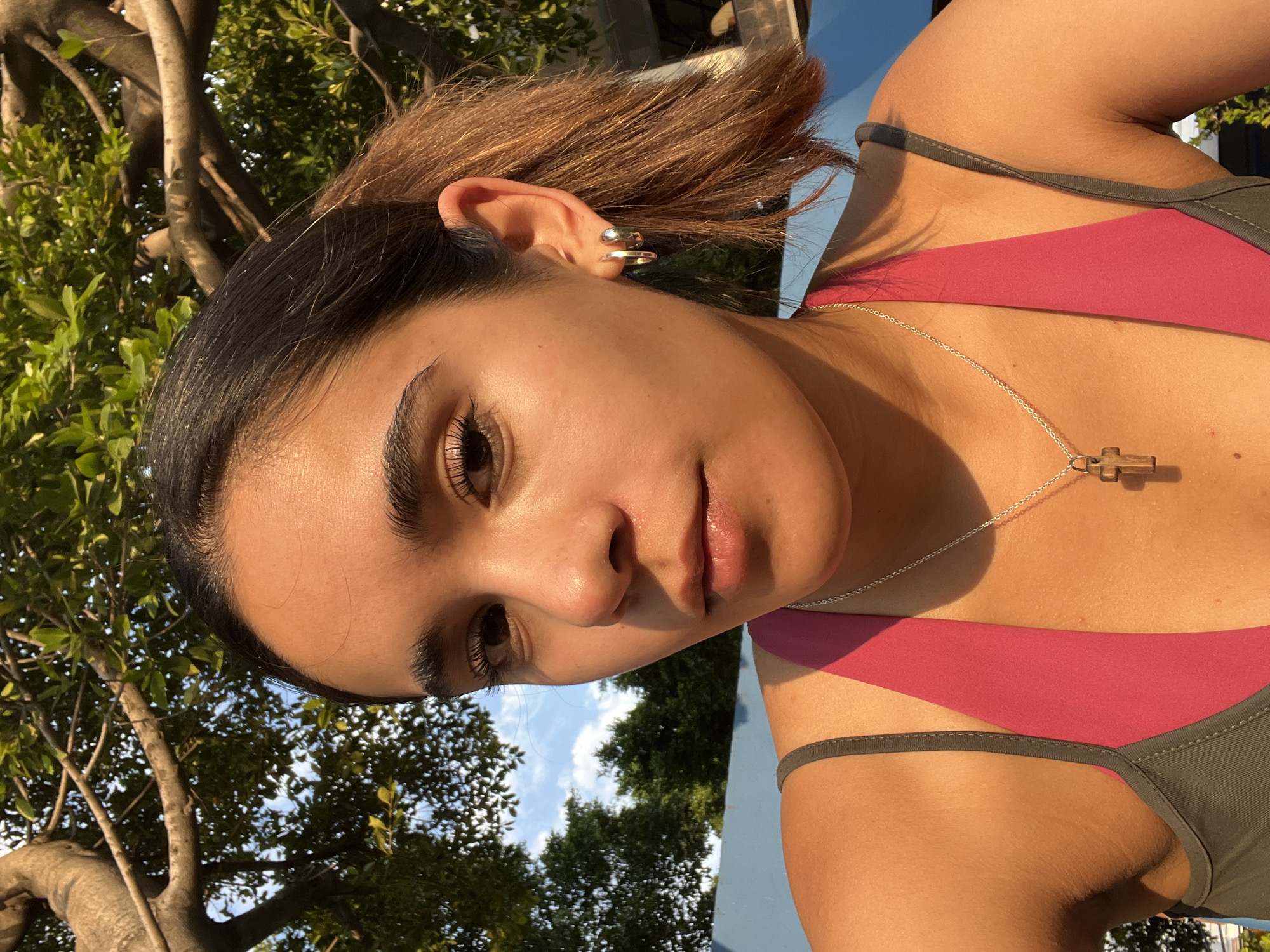 A medium shot shows a young woman against a backdrop of trees and sky, seemingly outdoors. The lighting suggests late afternoon or early morning. The woman is wearing a pink and dark green halter top. A silver necklace with a wooden cross pendant is visible. Her hair is short, dark at the roots, and light brown on the ends. She has multiple silver earrings on her left ear. The exact location in San Bernardino Tlaxcalancingo, Mexico, cannot be determined from the image alone.