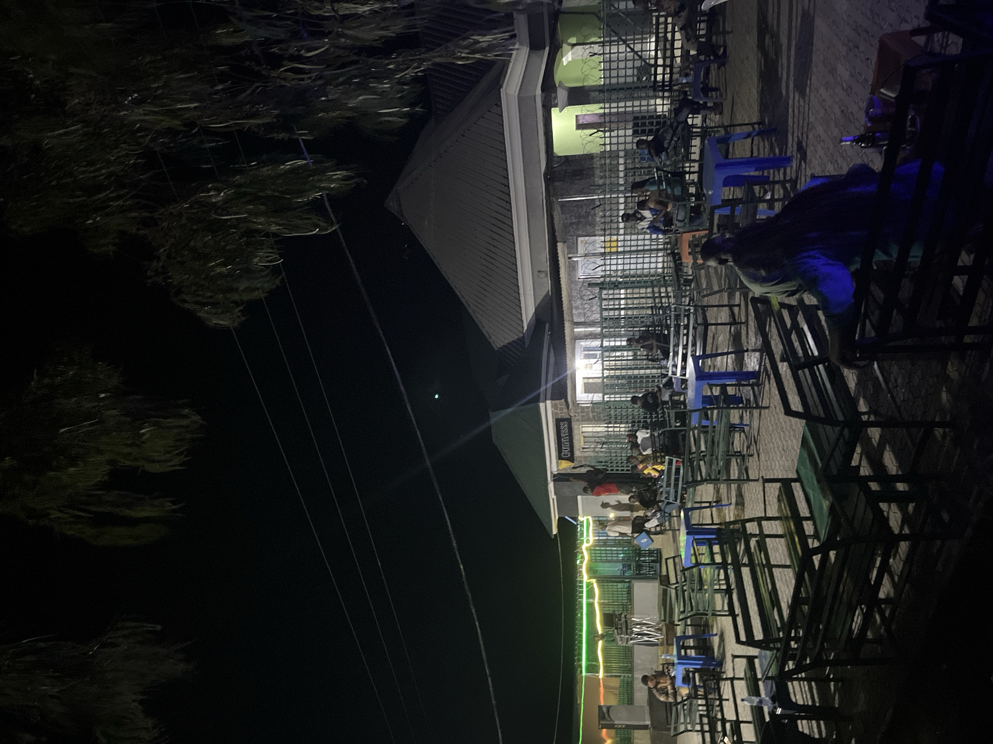 This image depicts an outdoor, nighttime social gathering space in Jalingo, Nigeria. The scene is illuminated by various artificial light sources, creating strong shadows and highlights.
The environment features a paved ground, likely interlocking stones, with numerous dark green benches and tables, some of which are blue plastic. Overhead, power lines traverse a dark sky, and the dark silhouettes of trees with long, drooping leaves are visible at the top edges of the frame.
Several buildings are present in the background. To the right, a building with a dark gray pyramidal roof and light green pillars/walls is visible behind a green grid-pattern metal fence. To the left of this, a smaller building features a prominent "Guinness" sign. Further to the left, another building or covered area is outlined with bright green and yellow neon lights, with a large dark speaker and a stand (likely for a DJ or performance equipment) positioned nearby. Green metal grid fences separate different sections of the area.
Multiple individuals are distributed throughout the seating area. In the foreground right, a person with dark hair, wearing a dark-colored top, sits on a bench with their back to the camera, with a bottle and other objects on the table in front of them. Towards the mid-ground left, a person in a brown or tan jacket sits at a blue table, appearing to focus on an object on the table. Other groups of people are seated and standing throughout the scene, notably near the "Guinness" sign, where a person in a red top stands, and further right, near the green-walled building. People appear to be socializing or relaxing.