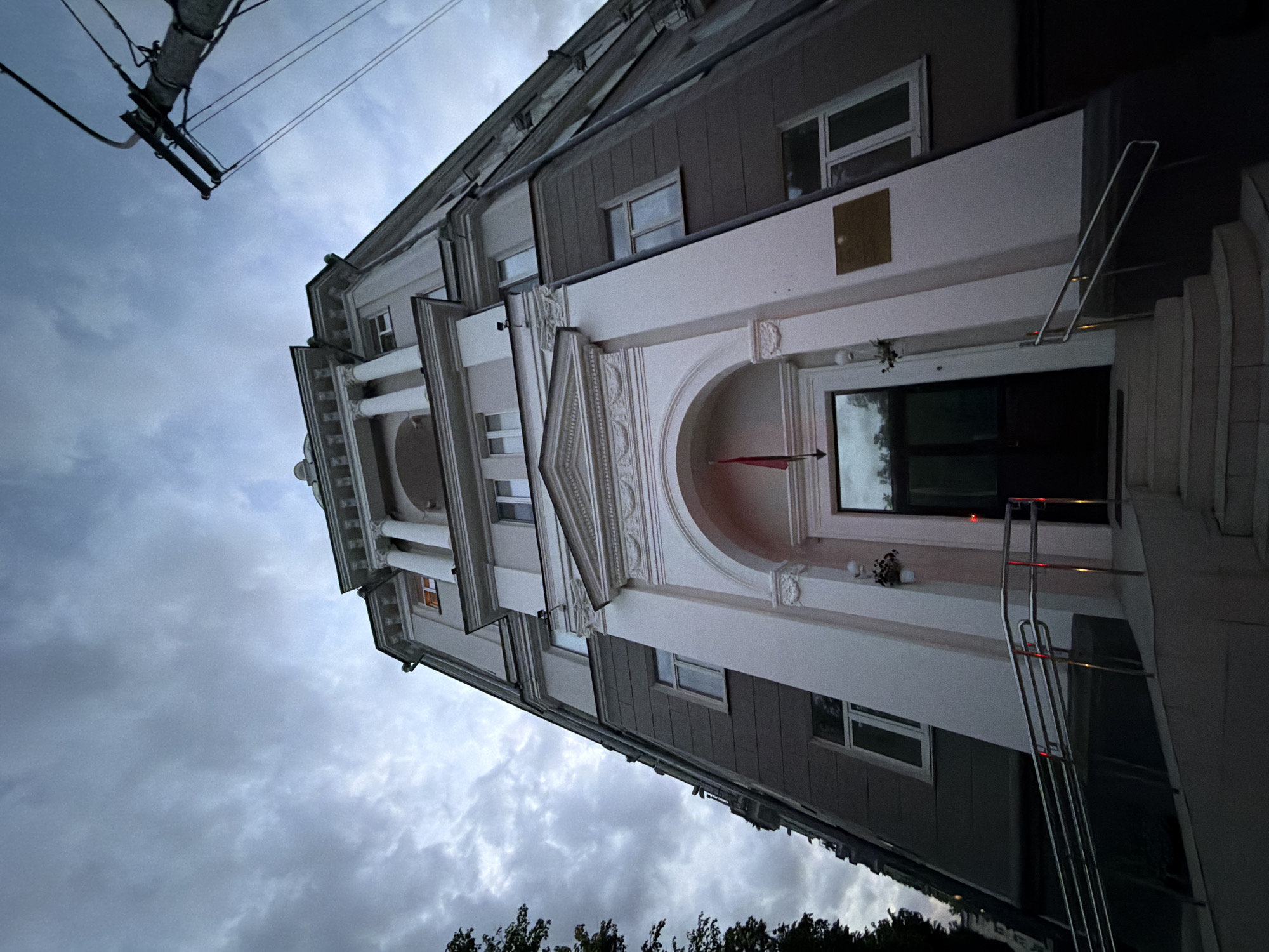 The image captures the facade of a stately building, possibly a government office or embassy, located in Minsk, Belarus. The architecture features neoclassical elements, including columns, arches, and decorative molding. The building's color scheme is predominantly gray and white. A flag is visible inside of the building. A chrome railing leads to the entrance. The sky is overcast with gray clouds, and a utility pole with wires is visible in the upper left corner. The building is situated in a urban environment, suggested by the trees at the bottom edge of the frame.