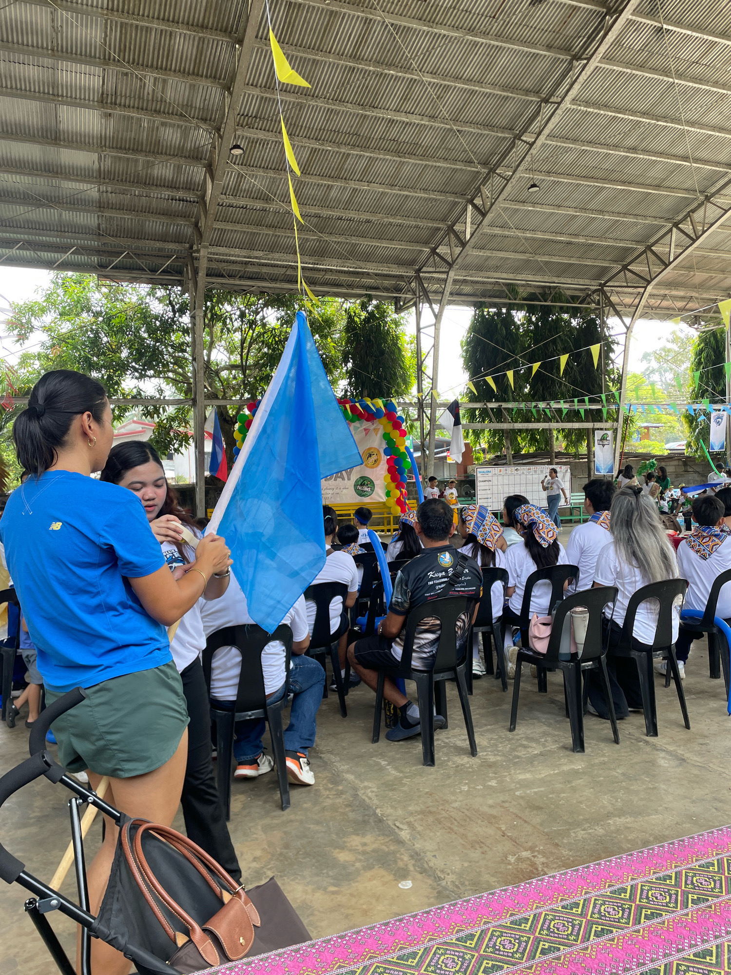 In Davao City, Philippines, an outdoor event is taking place under a corrugated metal roof supported by metal beams. The scene appears to be some sort of gathering or ceremony, possibly at a school or community center, given the presence of children and adults seated on black plastic chairs. In the foreground, two women are holding up a large blue and white flag. One woman, on the left, is wearing a blue t-shirt and green shorts, with her hair tied in a ponytail. The other woman is wearing a white shirt. Behind them, a crowd of people is seated, facing a stage area where a whiteboard or display board is visible. Balloons and banners, including one with "FALCONS" and a logo, decorate the background. Yellow triangular flags are strung across the roof. The ground is concrete, and a decorative pink and green carpet is visible in the lower right corner. The overall atmosphere seems festive and organized.