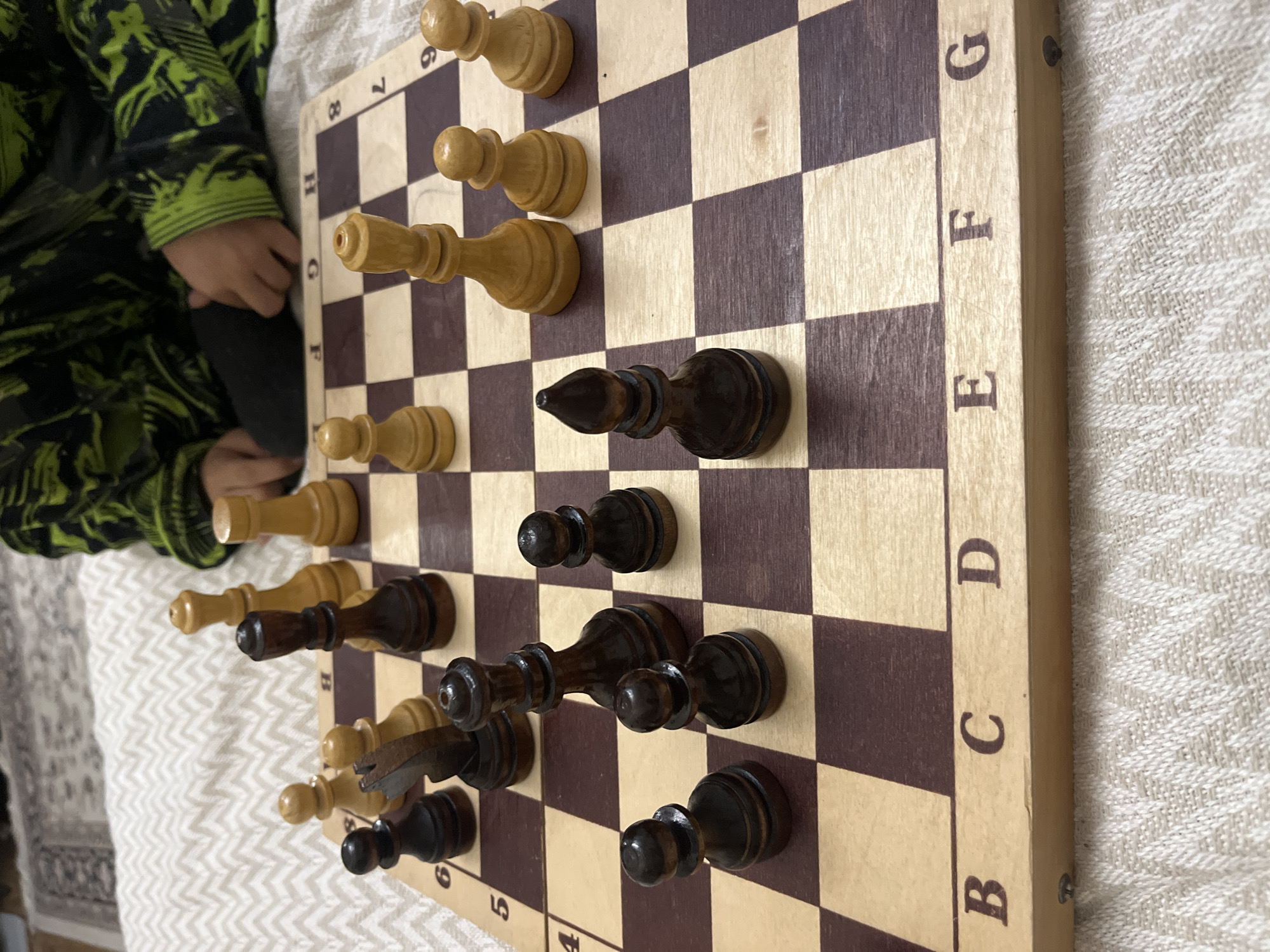 A top-down close-up shot shows a chessboard with chess pieces. The board is partially rotated. White pieces are on the top and bottom of the board. The black pieces are in the center. A child's hand is visible on the left edge of the image. The chessboard and the hand are resting on a patterned surface. There is no indication the image was taken in Kaspiysk, Russia.