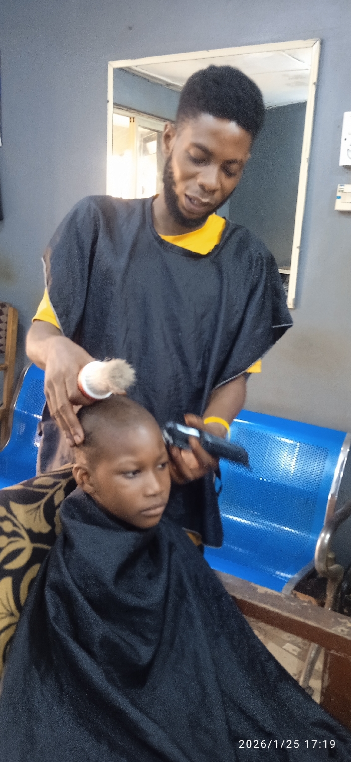 A barber with a beard and black hair is giving a child a haircut. The barber is wearing a yellow shirt and a black smock, and the child is covered with a black cape. The barber has one hand on the child’s head and the other holding a hair clipper. A shaving brush is placed on the child's head. They are in a barbershop, with a mirror and chairs visible in the background. The date "2026/1/25" is displayed in the lower right corner of the image.