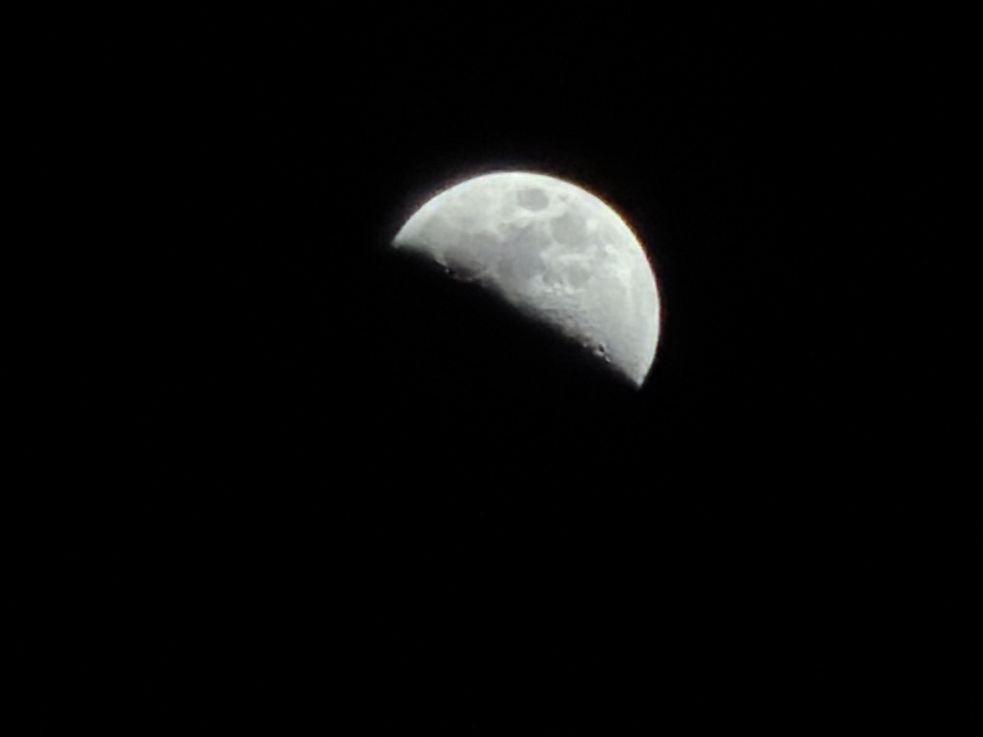 This media file features a clear, close-up shot of the Moon against a pitch-black night sky. The Moon is in a crescent phase, specifically appearing as a first quarter moon, with its right half brightly illuminated. The left side is in shadow, creating a distinct terminator line that marks the transition from light to dark.
Notable details of the lunar surface are visible, including several craters and smoother, darker areas known as maria. These features are clearly discernible on the illuminated portion of the Moon. The image captures the texture and irregularities of the lunar landscape. The edges of the Moon show a slight blur or chromatic aberration, suggesting the image might have been taken with a telephoto lens or through a telescope. The deep black background emphasizes the celestial body as the sole subject.
The setting is clearly nighttime. There are no other celestial bodies, clouds, or atmospheric disturbances visible, indicating a clear sky. No human activity or specific event is depicted other than the observation of the Moon. There is no visible text in the image. The location context for this image is Izhevsk, Russia.