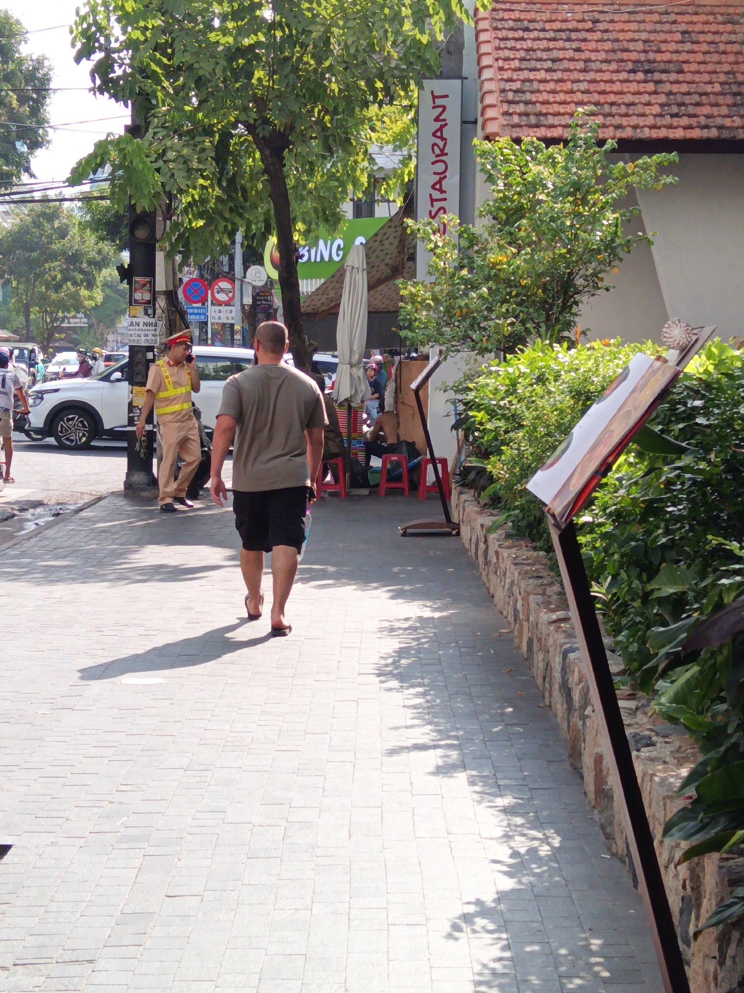 In Nha Trang, Vietnam, a man in a tan uniform and a reflective vest, possibly a traffic officer, stands on a sidewalk next to a traffic light pole and is speaking on a mobile phone. He is facing left and looking towards a white vehicle. A man in a grey t-shirt and black shorts walks away from the camera on the sidewalk. A street sign with Vietnamese text is visible on the traffic light pole. In the background, trees, a building with a tiled roof, and a sign that reads "RESTAURANT" are visible. The sidewalk is paved with light-colored tiles, and shadows from trees and an awning are cast on the ground. The overall scene depicts a sunny day on a city street.