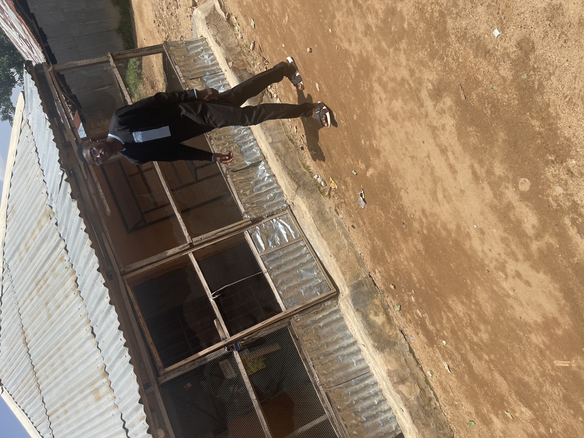 A person dressed in a black suit, white shirt, and black sandals stands in front of a structure with a corrugated metal roof. The person is facing the camera, with their body angled slightly to the left. They are wearing glasses and appear to be looking at the camera. The background consists of a dirt ground with some scattered debris. The scene is outdoors, under natural lighting, suggesting it is daytime. The overall atmosphere appears casual, possibly indicating a moment of pause or a posed photograph. The location is identified as Jos, Nigeria. There is no visible text in the image.