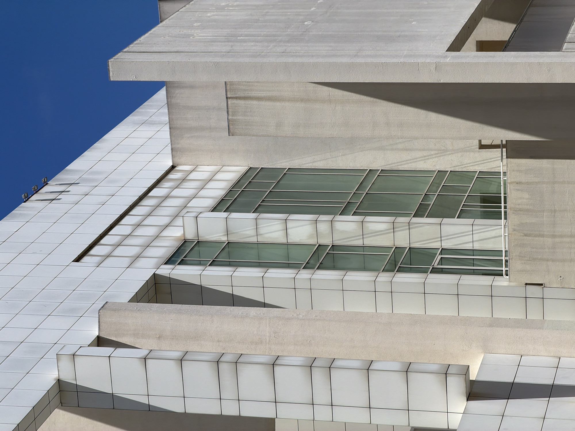 This is a photograph of a modern building exterior, likely a museum or cultural center, under a clear blue sky. The architecture is characterized by geometric shapes, clean lines, and a predominantly white and beige palette. The facade is composed of large white square tiles, some of which show signs of staining or weathering. Vertical beige pillars and sections of textured concrete punctuate the tiled surfaces.
A prominent feature is a large, multi-paned window with a green tint, divided by thin metal frames. The window wraps around a corner of the building. Shadows cast by the structure play across the surfaces, adding depth and definition. On the far right, a lighter colored concrete section is visible, with a recessed opening and what appears to be a railing.
The building's style is reminiscent of the architect Richard Meier's work, particularly his design for the Museum of Contemporary Art of Barcelona (MACBA). The clean, minimalist aesthetic, the use of white and geometric forms, and the prominent glass facade are all hallmarks of this style. Therefore, it is highly probable that this image depicts the MACBA, located in the El Raval neighborhood of Barcelona, Spain. There are no people or visible signs of activity in this static architectural study.