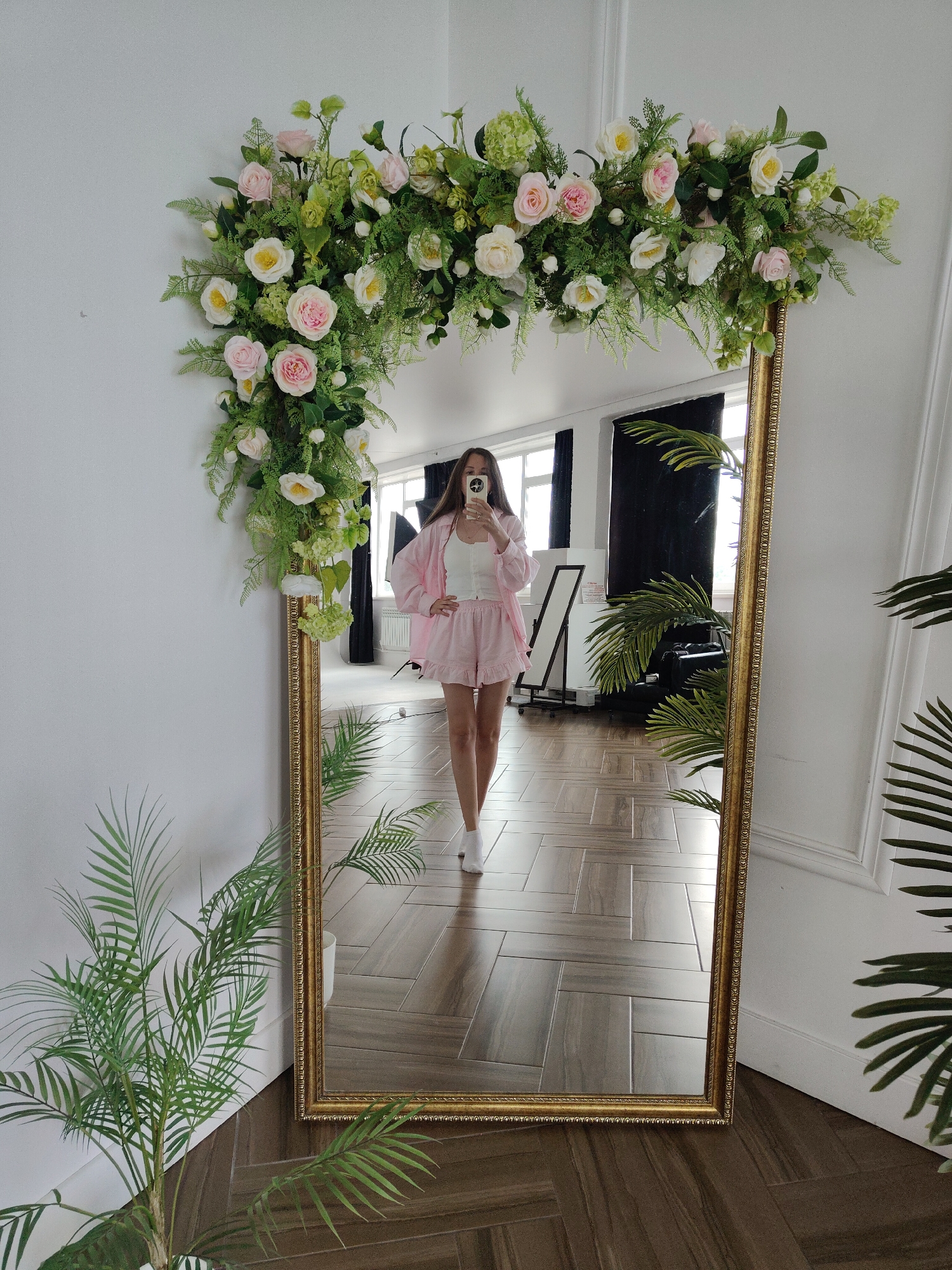 A full-length mirror with a gold ornate frame stands against a white wall, adorned with a large floral arrangement of roses and greenery. The mirror reflects a young woman in pink shorts and a white top, holding a phone to take a selfie. She is standing in a room with wooden floors and large windows. To the left of the mirror, a potted plant with green fronds is visible. The room appears to be well-lit with natural light coming from the windows.