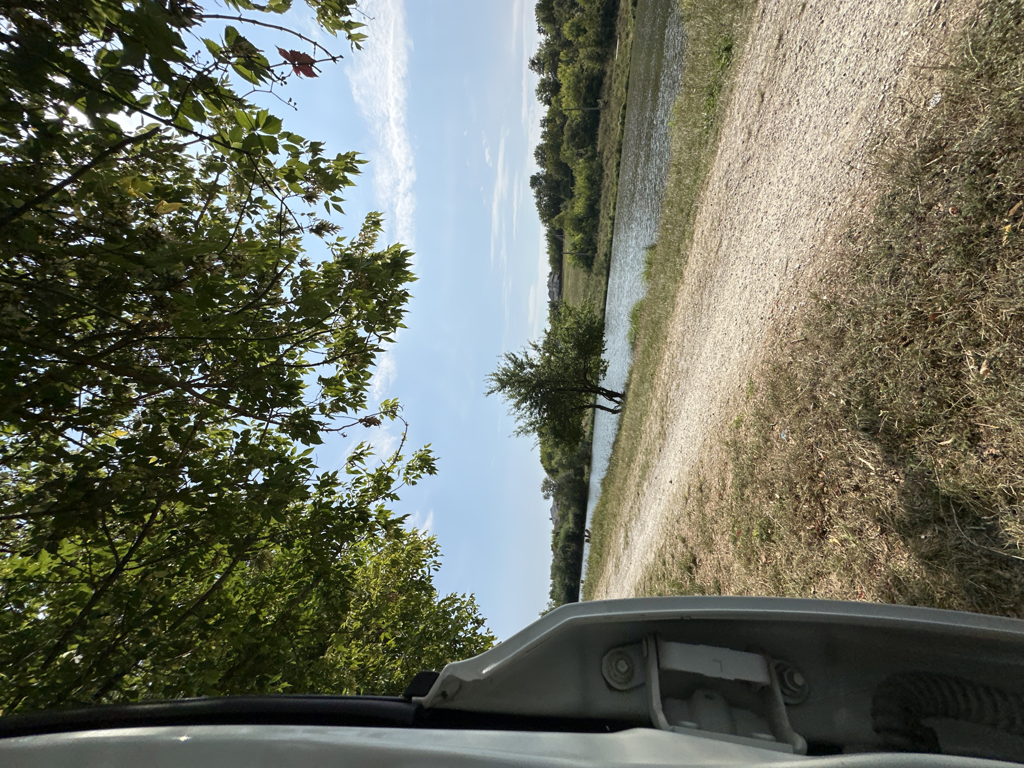 The image shows a serene landscape beside a body of water, with a gravel path leading into the distance. The foreground is dominated by the partially opened door of a light-colored vehicle, revealing its interior hinge mechanism. Lush green trees with dense foliage overhang the scene from the left and top, with sunlight filtering through the leaves. Beyond the path, a calm river flows, bordered by a dense line of green trees and vegetation. In the background, a few houses are visible, suggesting a rural or semi-rural setting. The sky is a clear blue with scattered white clouds. The environment appears to be a park or natural area, possibly near a settlement in Makiv, Ukraine. There are no people or animals visible in the frame. The overall atmosphere is peaceful and natural.
