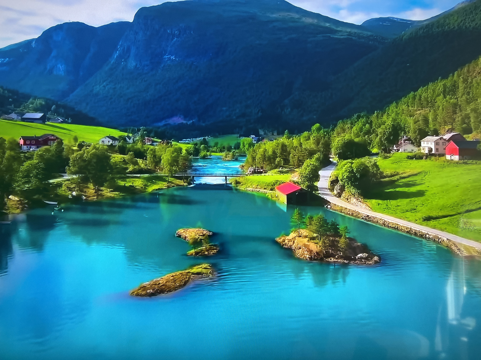 The image displays a scenic landscape featuring a vibrant turquoise lake with several small, rocky islands. Lush green meadows and forests flank the water, contrasting with the dark blue, mountainous backdrop. A few houses with red roofs dot the landscape. A bridge spans a narrow part of the lake, and a road runs alongside the water's edge.
This scene does NOT appear to be located in Basingstoke and Deane, United Kingdom. The landscape is consistent with the fjords found in Norway.