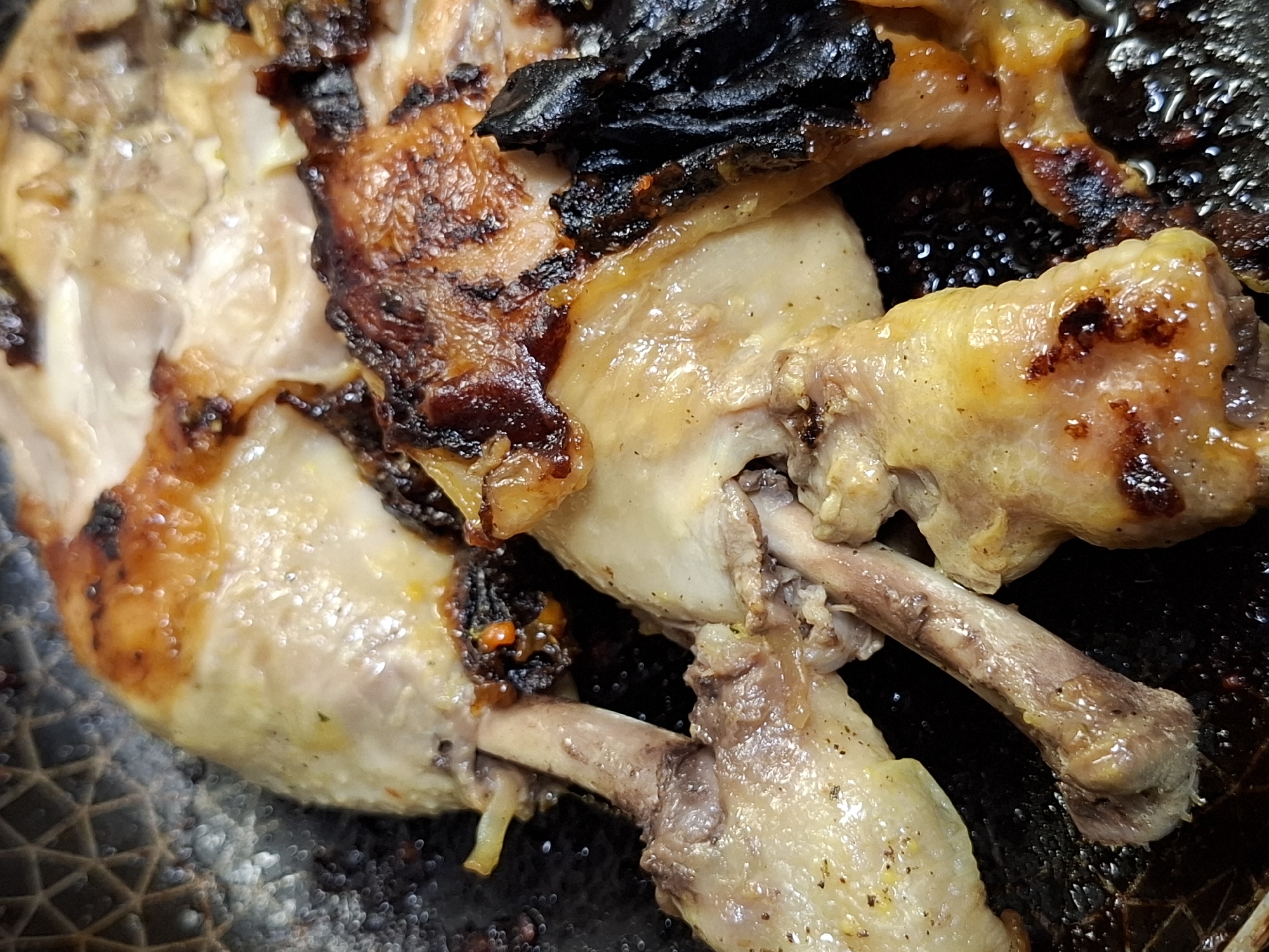 The image shows a close-up of cooked chicken pieces in a dark, likely pan. The chicken has a mixed appearance, with some parts appearing golden brown and well-cooked, while others are charred or blackened. The bones are visible on some pieces of the chicken, with a shiny texture suggesting moisture. The background of the pan is dark and oily. There's no evidence suggesting the location is Novi Sad, Serbia; the setting appears to be a kitchen or cooking environment. No people are visible. The image primarily displays food.