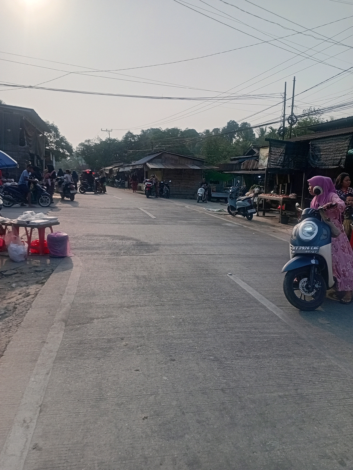The image depicts a street scene in Anggana, Indonesia, likely a market area. A paved road runs through the center of the frame, with various buildings and stalls lining both sides. Several motorcycles are parked or being ridden by people. On the right side of the road, a woman wearing a pink hijab and a pink patterned dress is standing next to a dark blue and white scooter. Another woman and a child are standing nearby. On the left side of the road, a stall is set up with red plastic stools and bags of items. The sky is bright, suggesting daytime, and power lines are visible overhead. The overall impression is a busy, informal commercial environment in a tropical setting.