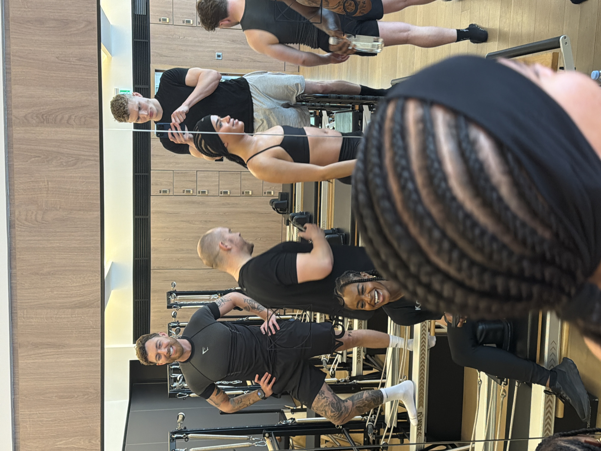 The image depicts a group of people exercising in what appears to be a Pilates or fitness studio. The studio has multiple Pilates reformers, and a wall of wooden lockers. The scene is reflected in a large mirror that is visible in the background. The reflection shows five people, some of whom are using the Pilates reformers. One person, in the foreground, is smiling and appears to be interacting with the camera. The overall impression is of a modern, well-lit fitness facility in London.