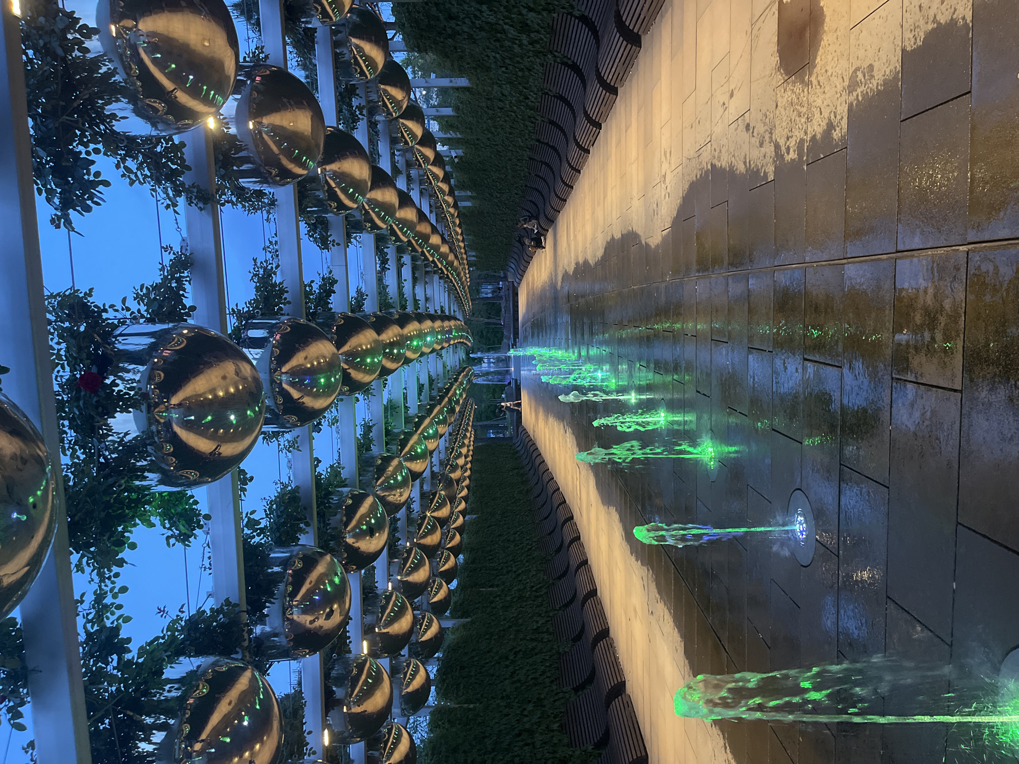 The image shows a water feature and art installation in Krasnodar, Russia, at night or dusk. On the left, a row of reflective spheres hangs in a linear arrangement, interspersed with greenery. To the right, a dark tiled wall has jets of water with green lighting shooting out horizontally. A walkway runs parallel to the wall. Two people are seen on the walkway further in the distance.