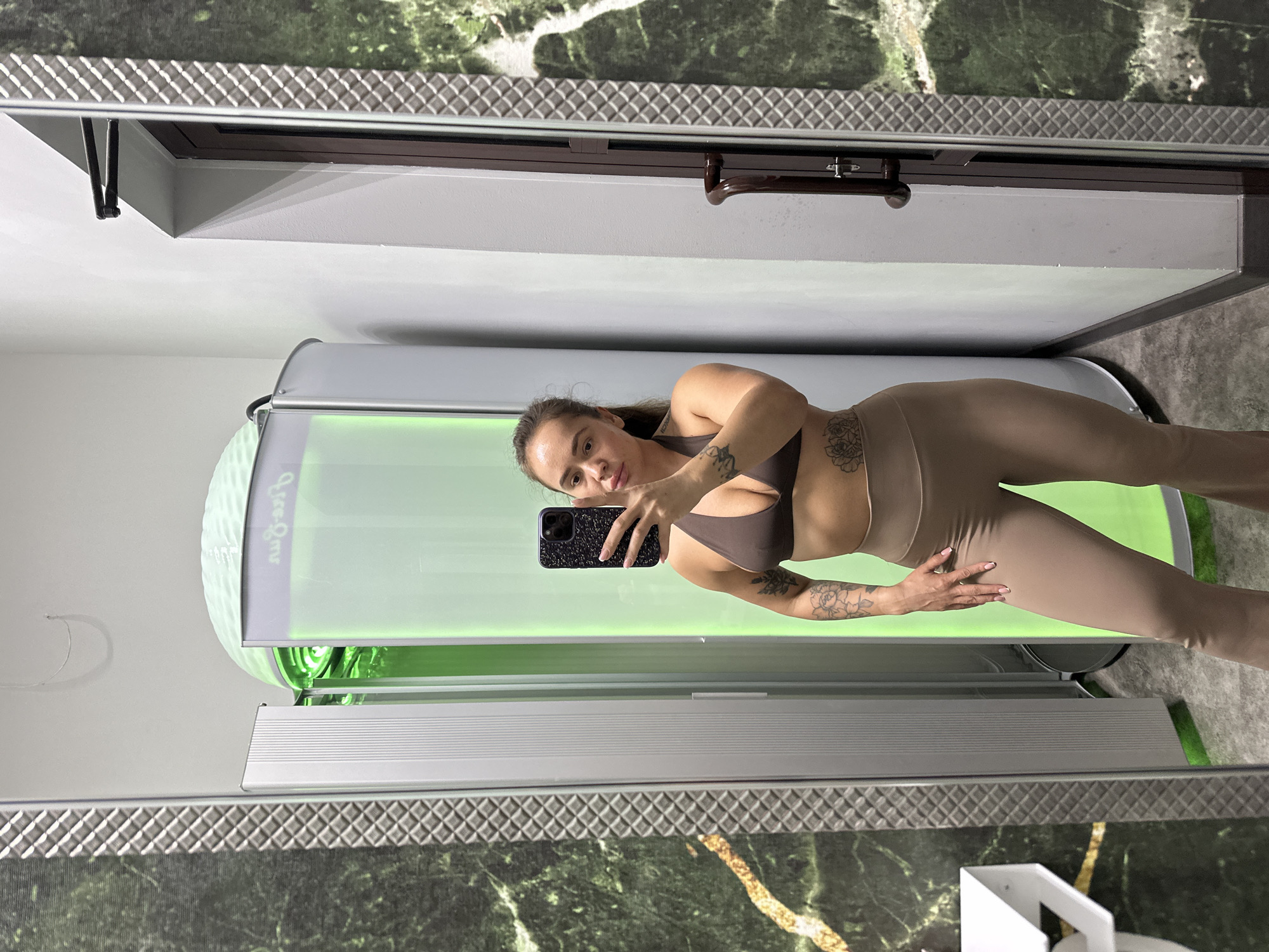 A woman in athletic wear is taking a selfie in a tanning bed. She is wearing a brown sports bra and taupe leggings. Her hair is pulled back in a ponytail, and she has tattoos on her arm and thigh. The tanning bed has a bright green interior and a silver exterior. The walls of the room are covered in a dark green marbled wallpaper. The floor appears to be a speckled grey. The lighting in the room is bright, suggesting it might be daytime or the tanning bed lights are on. The woman's expression is neutral. Visible text on the tanning bed reads "Gee-Goo".