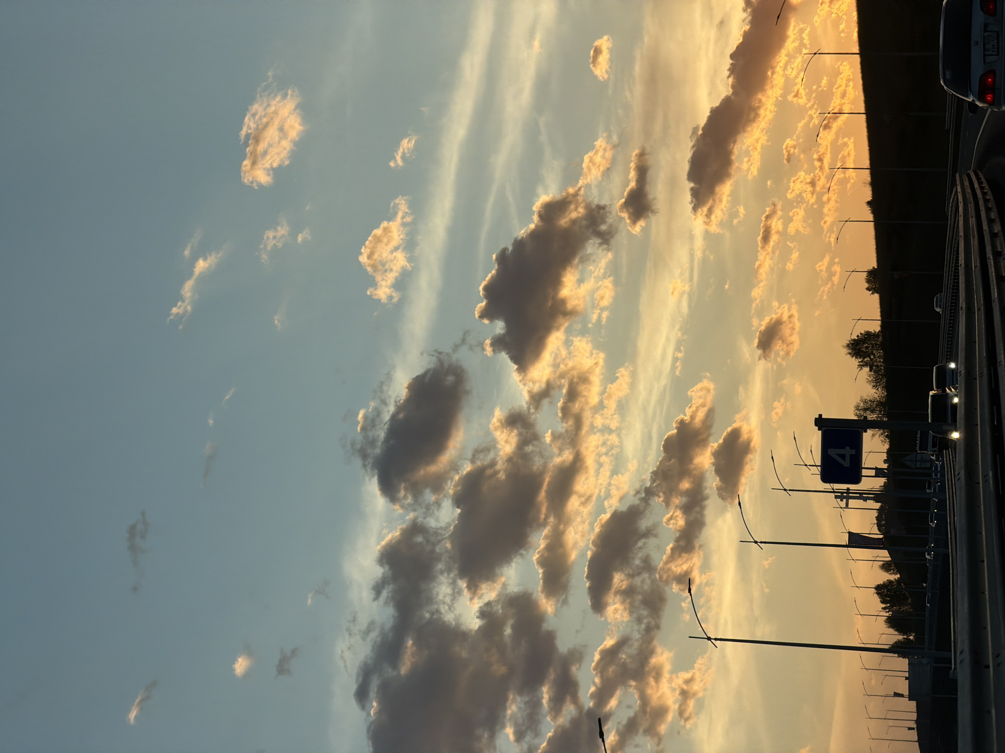 The image shows a sunset sky in Krasnoyarsk, Russia, seen from a highway. The sky is a mix of blue and orange hues, with clouds illuminated by the setting sun. A highway runs along the bottom of the frame, with cars and lampposts visible. A road sign with the number "4" can be seen along the highway. The scene depicts a typical evening on a highway in Krasnoyarsk, with traffic moving against the backdrop of a colorful sunset.