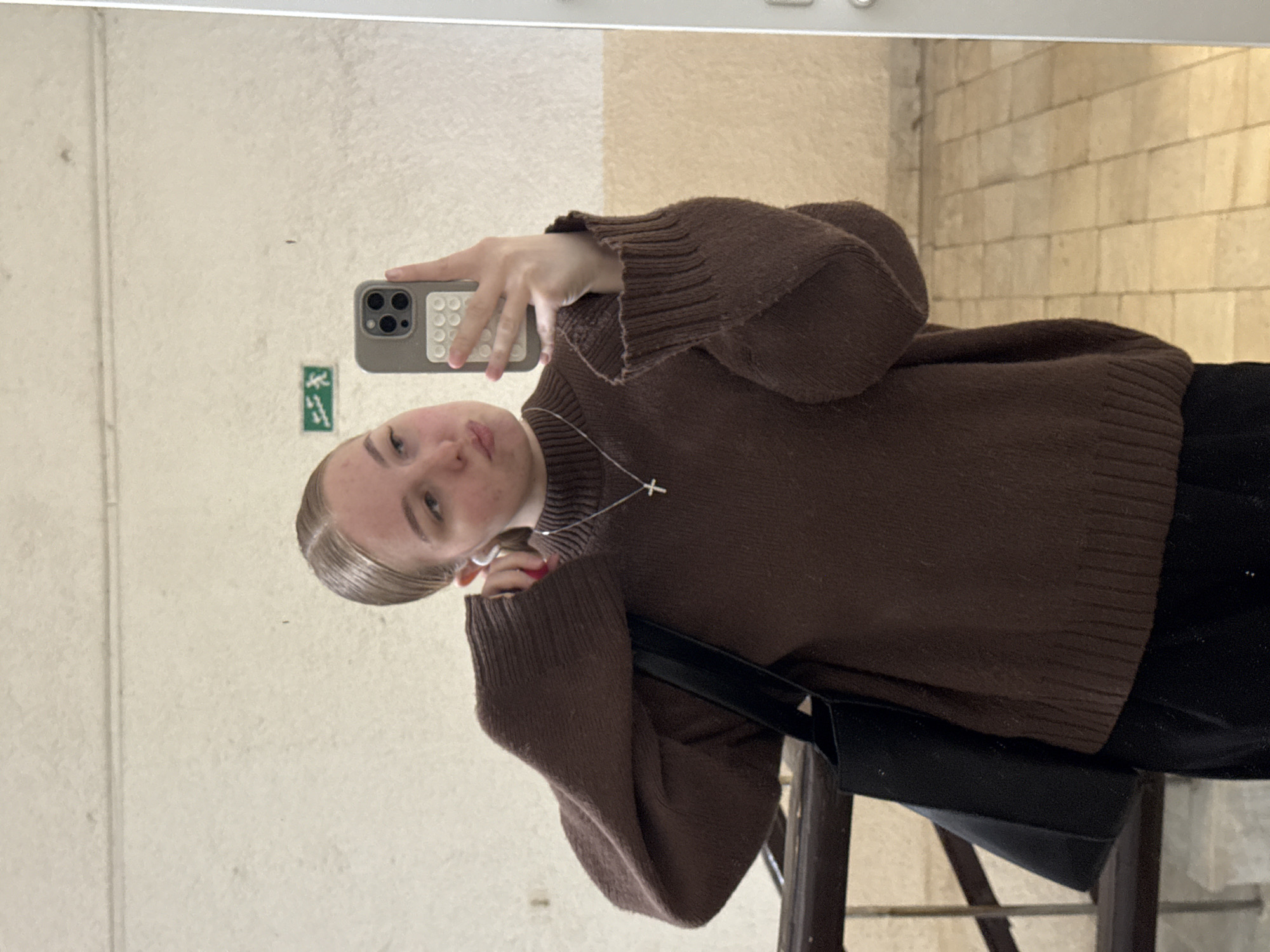 A young woman is taking a selfie in a mirror. She is wearing a brown turtleneck sweater and a necklace with a cross pendant. She is holding a smartphone with a light-colored case in her right hand. The background appears to be an interior space with light-colored walls, possibly a public building. There is an emergency exit sign on the wall behind her. The location is likely Perm, Russia. The selfie seems to be taken casually.