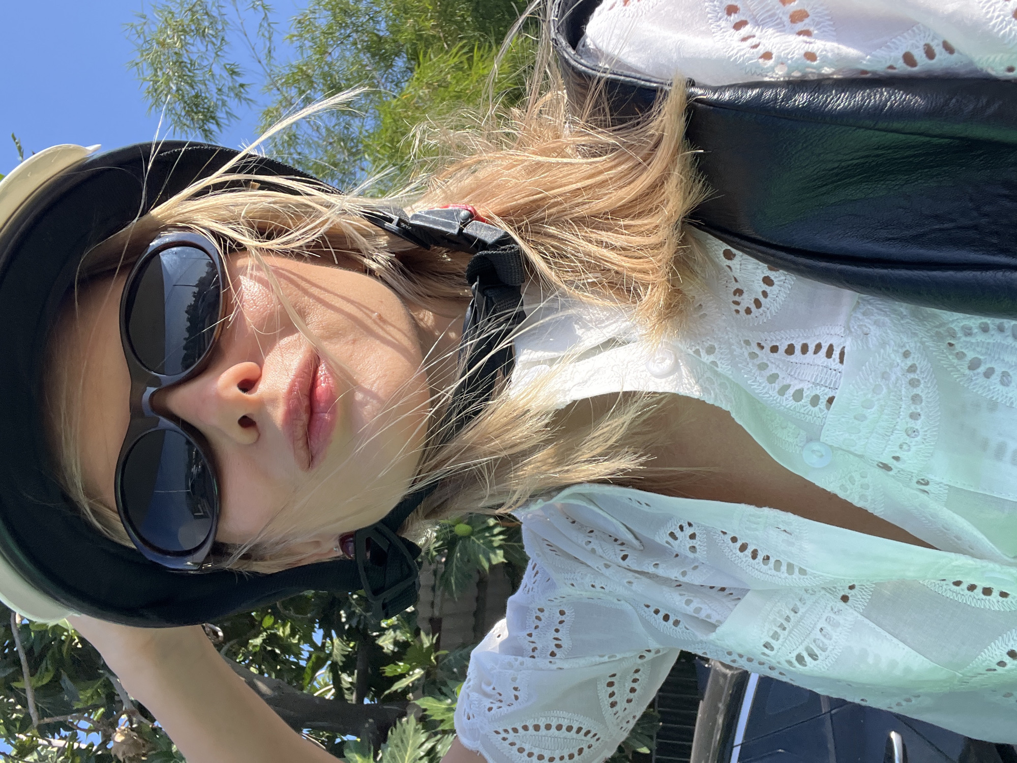 A woman with blonde hair and sunglasses is wearing a white eyelet shirt and a helmet, and holding her hand to the helmet. She is outdoors, with trees and a clear blue sky in the background. The image appears to be a selfie, taken from a close-up perspective. There is no visible indication of the city of Nha Trang Ward, Vietnam.