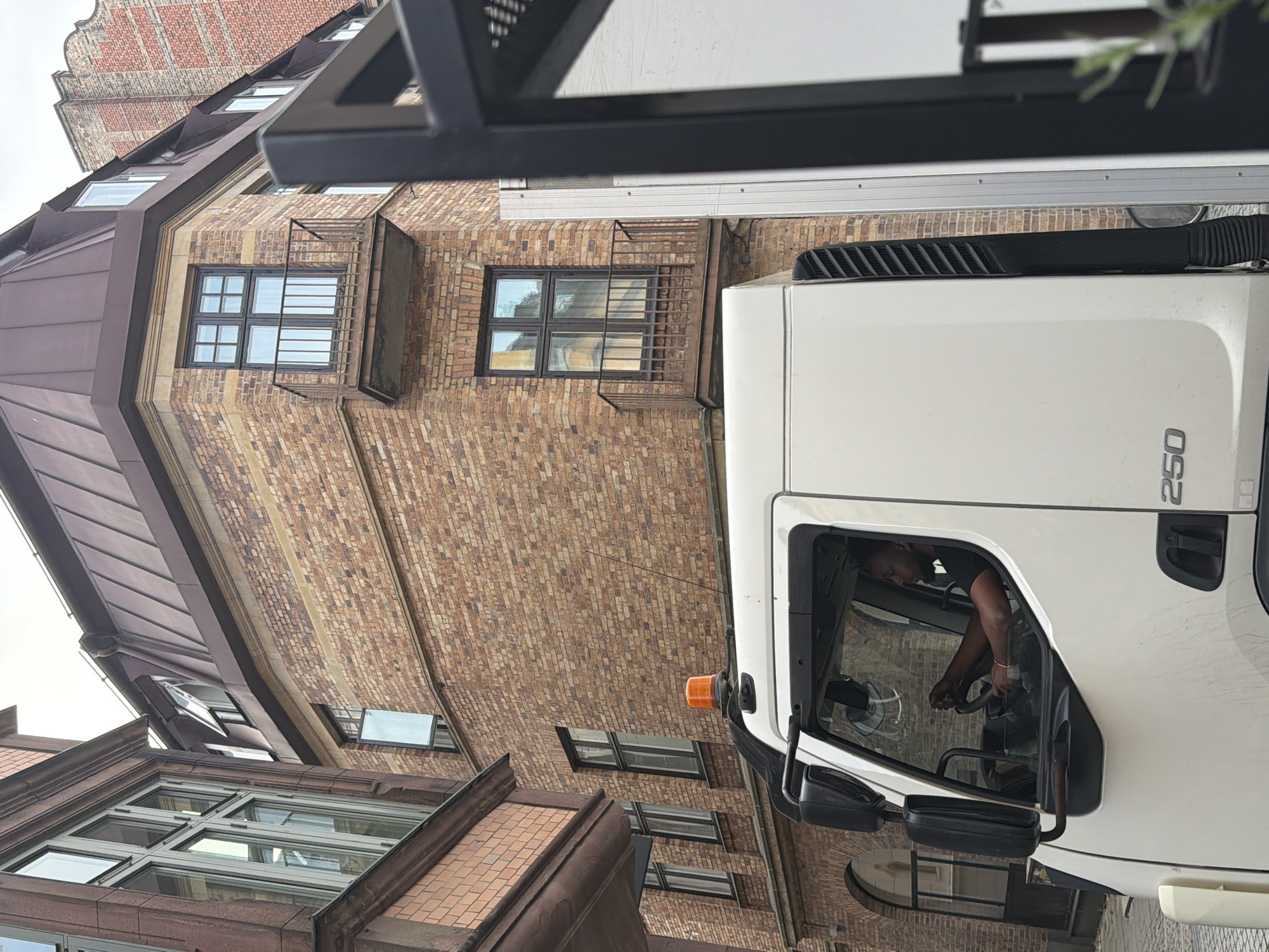 In the city of Malmö, Sweden, a white truck with an orange safety beacon on its roof is parked on a cobblestone street. The truck has the number "250" visible on its side. A Black male is seated in the driver's seat, holding the steering wheel. The truck is positioned in front of a multi-story brick building with several windows and balconies. To the left of the brick building, another building with red brick and large windows is visible. A dark metal fence is in the foreground on the right side of the image, partially obscuring the truck and the building behind it.