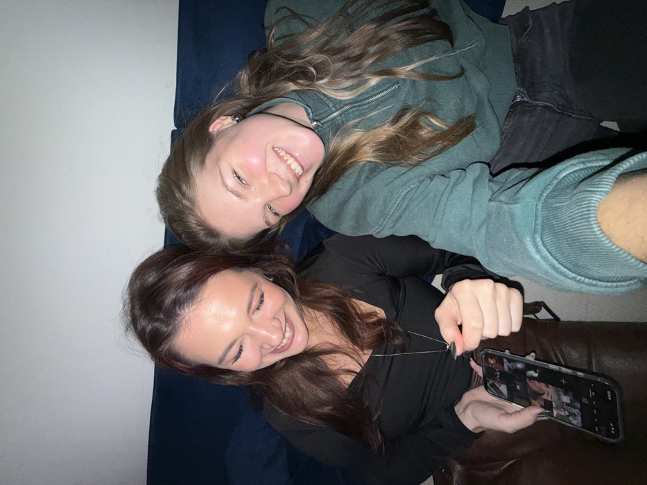 Two smiling women lie side-by-side on a couch. One woman with blonde hair wears a green sweatshirt and jeans. The other woman with reddish-brown hair wears a black top and holds a phone displaying a collage of images. They both appear relaxed and happy. The couch has a blue cushion on one side and brown leather on the other. The background is a plain white wall.