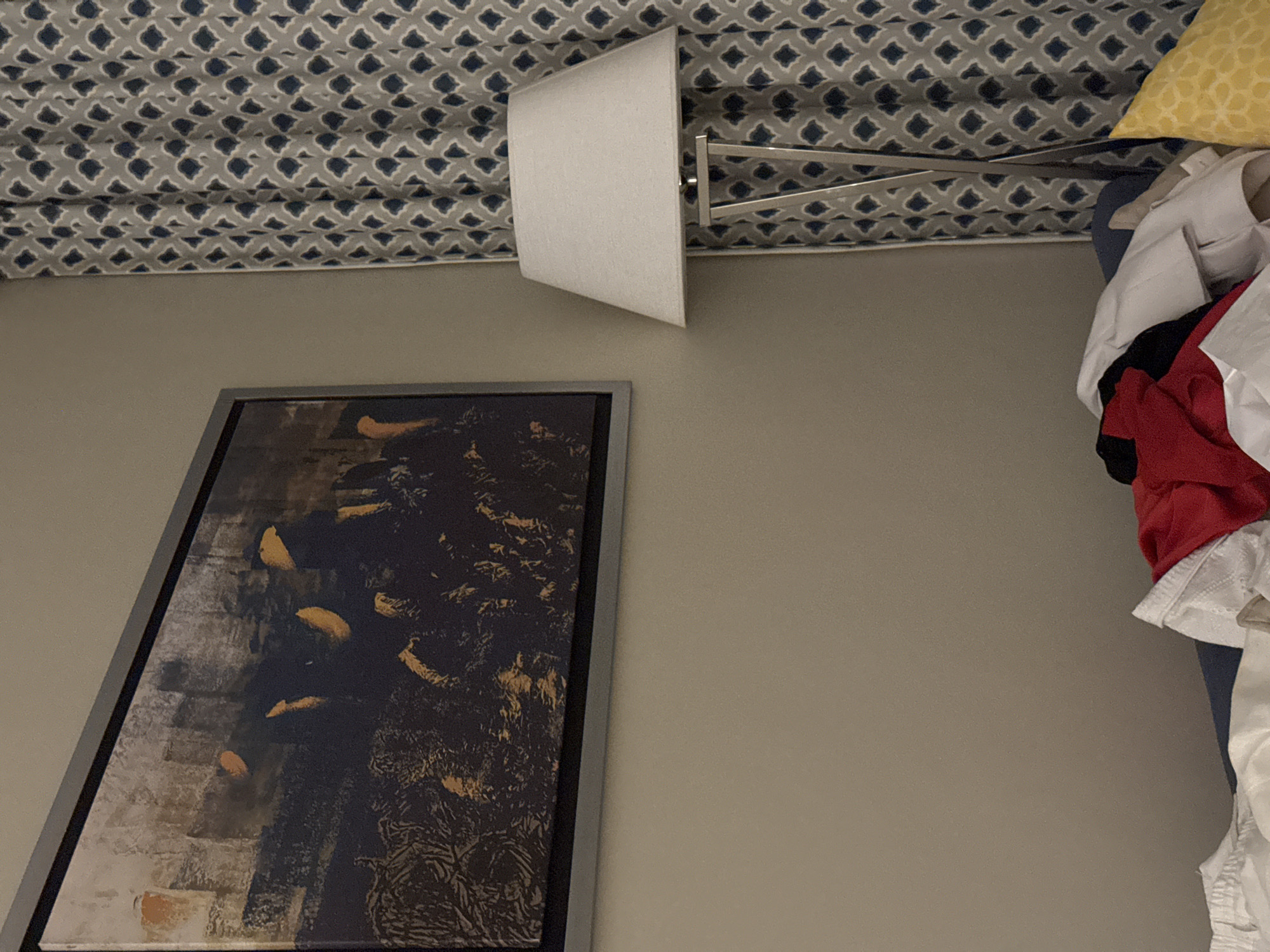 A floor lamp with a white conical lampshade stands next to a patterned curtain in a dimly lit room. The curtain has a repeating diamond pattern in blue and white. To the left of the lamp, a framed abstract painting hangs on a neutral-colored wall. The painting features dark tones with strokes of orange and yellow. In the foreground, a pile of clothes, including a red shirt, lies on a surface, possibly a bed. A yellow pillow with a geometric pattern is visible on the right. The room appears to be a bedroom or living area.