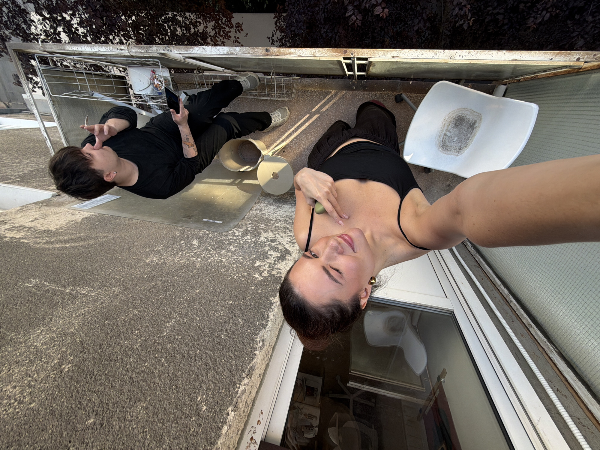 The image depicts two individuals on a concrete balcony. A person, likely a male, is lying on their back, holding a cigarette in their mouth and a phone in their right hand. They are wearing a long-sleeved black shirt and dark pants. There's a bucket nearby. Another person, likely female, is taking a selfie; she has her arm outstretched, pointing towards her chest, and is wearing a black top with spaghetti straps. A white plastic chair is near her, and a window with items inside is visible in the background. The balcony has a metal railing and seems to be on an elevated level. The location could be Belgrade, Serbia.