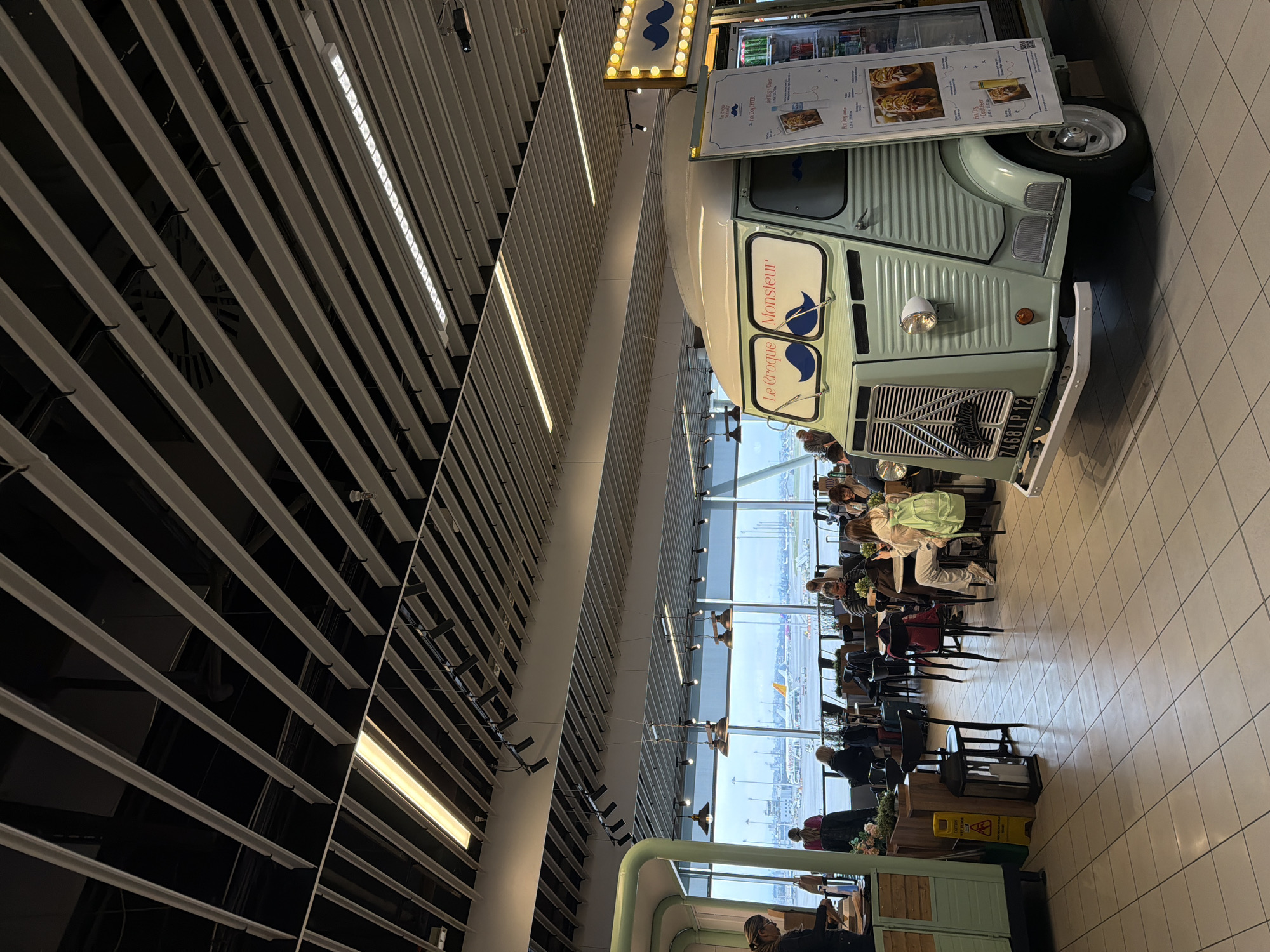 The image shows a food truck named "Le Croque Monsieur" parked inside what appears to be an airport terminal. Passengers are seated at tables nearby, eating or drinking, with a view of the airfield through large windows. The ceiling features a modern design with linear lighting fixtures. The food truck's license plate reads "7468 LP 12." Food and drink options are displayed on a menu board attached to the truck. The location is likely the Sofia Airport in Sofia, Bulgaria.