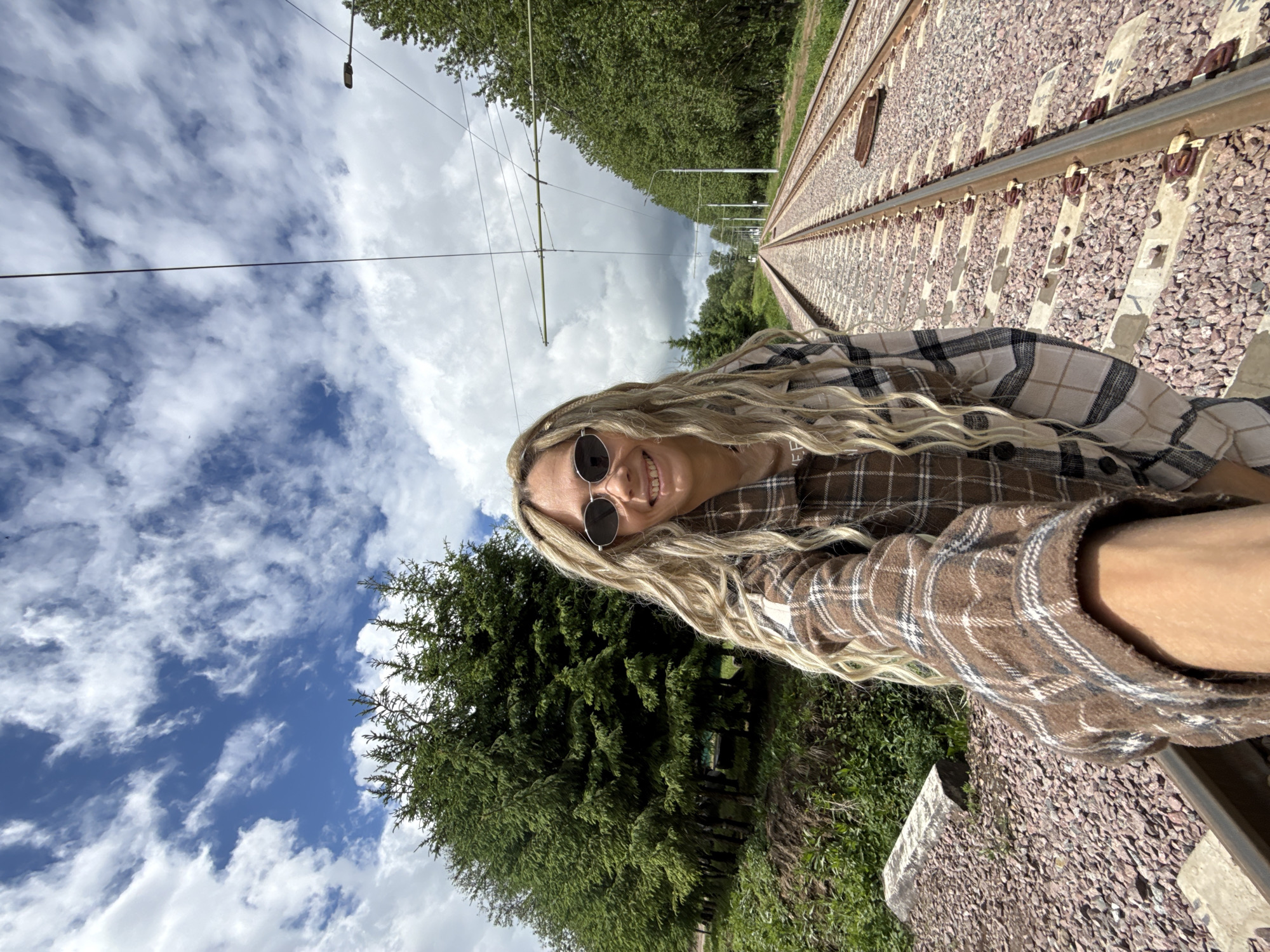A selfie of a woman with blonde hair and sunglasses standing on a railway track. She is wearing a brown and white plaid shirt. The railway track extends into the distance. Vegetation is visible on either side of the track. The sky is blue with white clouds. The location appears to be Rusanovo, Russia.
