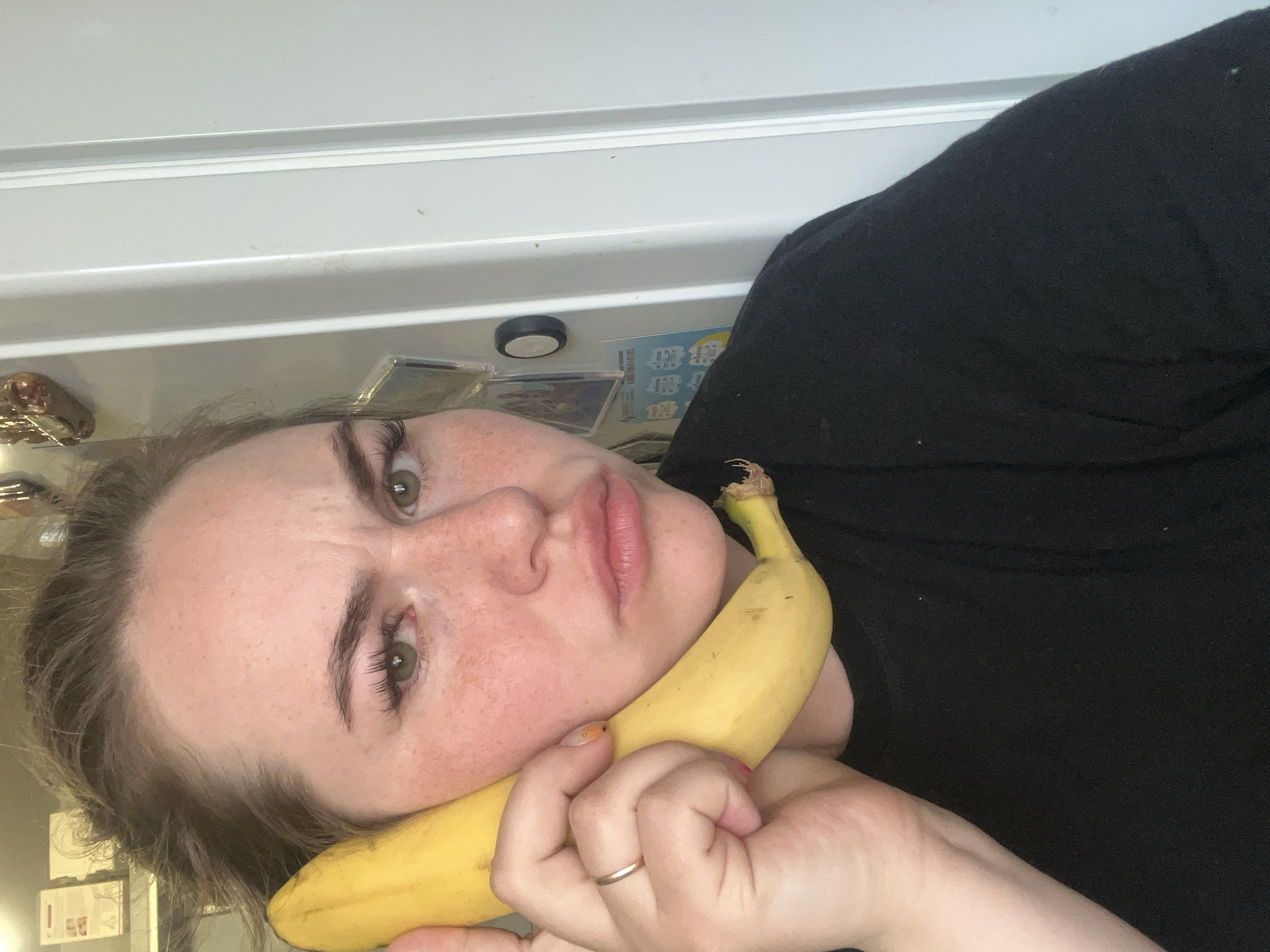 An overhead, rotated image captures a young person (likely female) looking directly into the camera with a neutral, slightly unamused expression, featuring a subtle pout and a raised left eyebrow. She has light brown hair, prominent freckles across her nose and cheeks, and green or hazel eyes. She is wearing a plain black top.
The central activity involves the person holding a ripe yellow banana against her left cheek and mouth. Her left hand, adorned with a simple gold-colored ring on the ring finger and bright orange nail polish, secures the banana.
The background consists of a large white surface, suggestive of a refrigerator or white cabinetry in a domestic setting. Attached to this surface are a small black circular object (possibly a magnet) and several small, square items. One of these square items displays indistinct, blurred text that appears to be non-Latin characters. The lighting is consistent with an indoor environment, offering no clues about the time of day or outdoor weather. The person's posture and the camera angle suggest a casual, spontaneous moment. The location context for this scene is Chaikovsky, Russia.