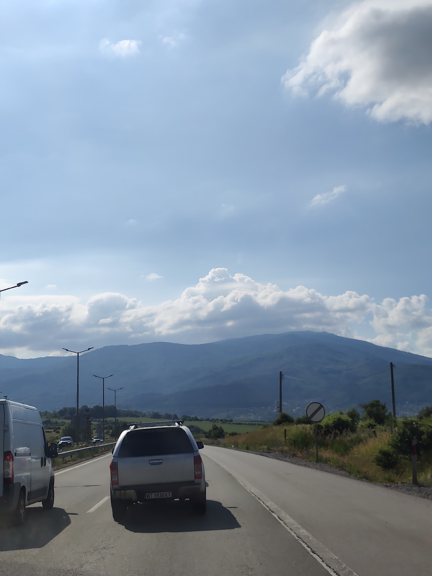 The image depicts a view from a vehicle traveling on a highway. In the foreground, a silver pickup truck with a camper shell is visible, displaying the license plate "BT 1930 KT". To the left, the rear of a white van is partially visible. The road ahead curves to the right, with a speed limit sign indicating the end of any restrictions. The landscape features rolling green hills and mountains in the background, with scattered trees and vegetation along the roadside. The sky is a light blue with scattered white and gray clouds. Streetlights are visible along the highway, suggesting it is a developed area. The presence of the license plate suggests a location in Bulgaria. Based on the scenery and the specific license plate format, the location is likely near Golemo Buchino, Bulgaria.