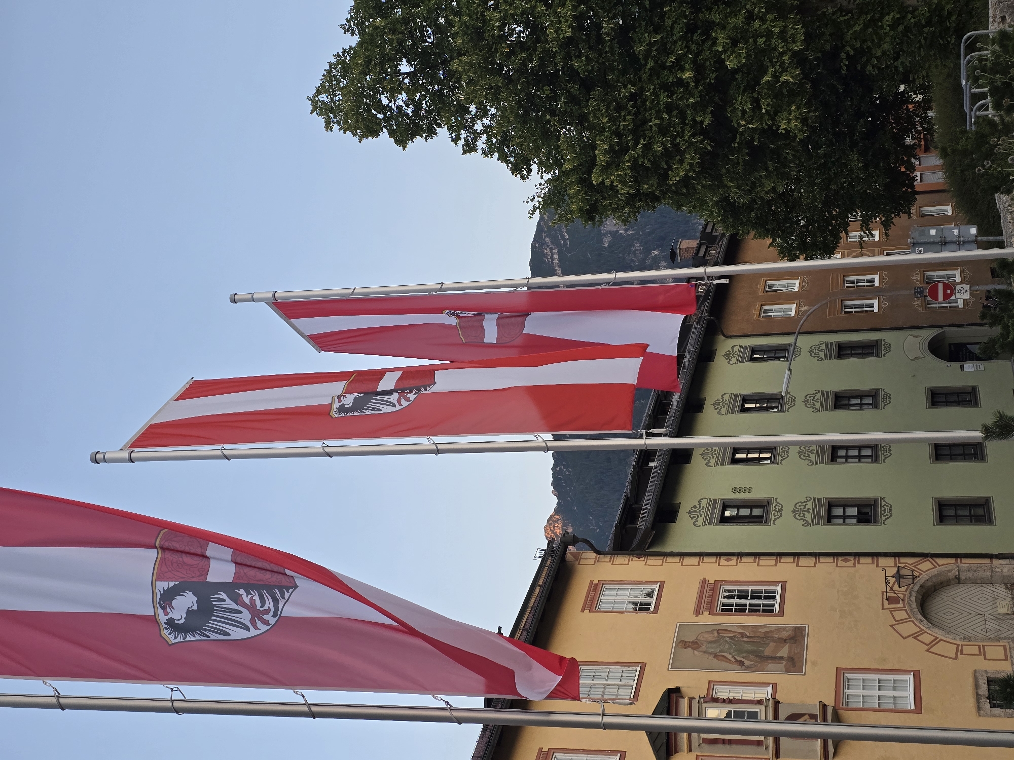 The image captures a street scene in Garmisch-Partenkirchen, Germany, featuring several tall flags with red and white stripes, each bearing a coat of arms. In the foreground, three flags are prominently displayed, obscuring parts of the buildings behind them. To the left, a yellow building with multiple windows and a decorative archway is visible, featuring a mural of a man. Adjacent to it is a light green building with decorative patterns around its windows. Further to the right, a brown building with white-framed windows and a large, leafy tree fill the frame. In the background, a mountain range is visible, with the sky a clear, pale blue. The scene appears to be set during daylight hours, possibly late afternoon, given the soft lighting.
