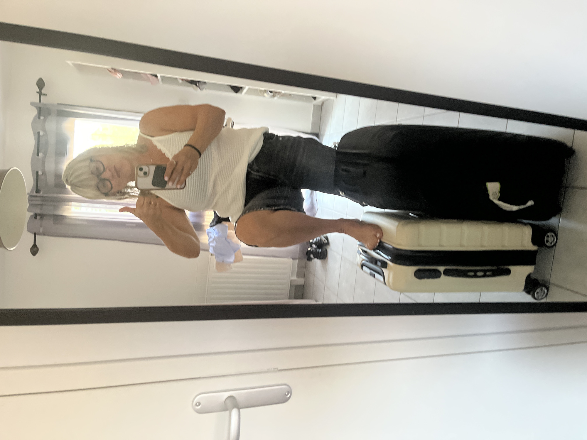A blonde woman with glasses and a white tank top is taking a selfie in a mirror. She is standing on top of two suitcases, one light-colored and one black, with her right foot on the light-colored suitcase and her left leg bent over the black suitcase. She is holding a phone in her right hand and giving a thumbs up with her left hand. The background appears to be a room with a window, a radiator, and some clothing on the floor. The location is specified as Chambœuf, France.