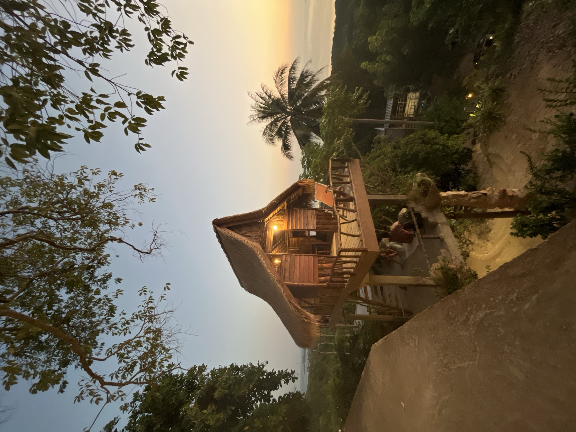 A wooden hut with a thatched roof is perched on stilts on a hillside overlooking the sea at sunset. The hut has a balcony with two orange chairs and a wooden railing. Several people are seated on the ground below the hut. The scene is located in Karimunjawa, Indonesia. Palm trees and lush green vegetation surround the hut. The sky is a gradient of pale blue to warm orange, indicating the setting sun. A paved path is visible in the foreground.