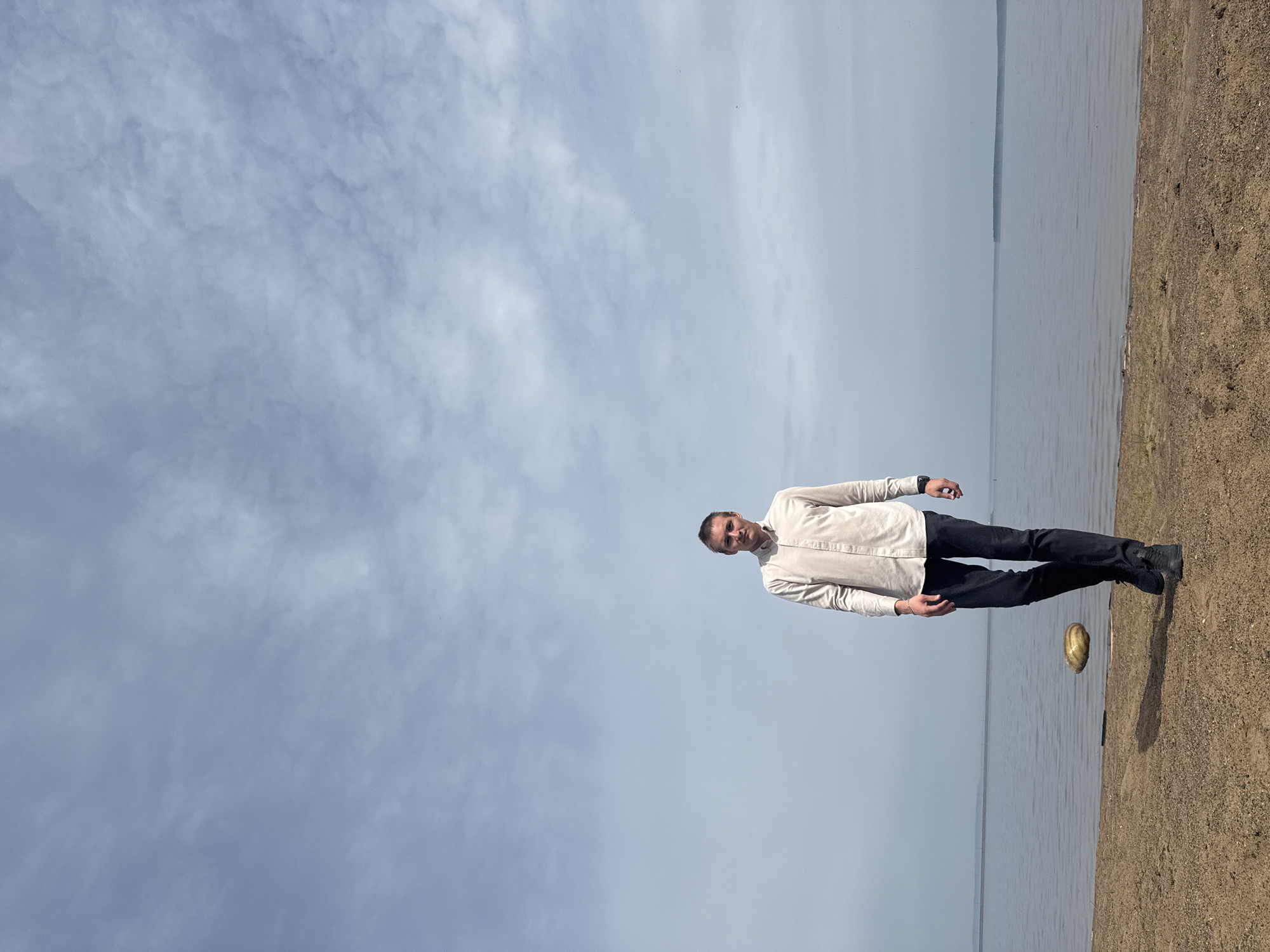 The image depicts a man standing on a beach in Chaikovsky, Russia. He is wearing a light-colored shirt and dark pants. The beach is sandy and the sky is cloudy. The water is calm. The photo appears to be taken from a low angle. There is an object (possibly a shell) on the ground.