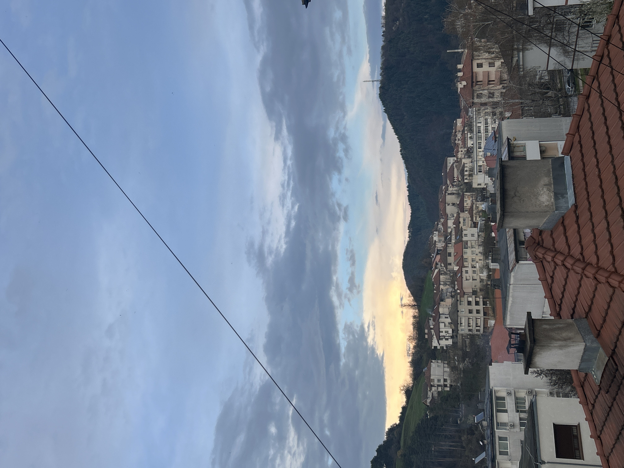 The image captures a landscape view in Zlatograd, Bulgaria. The sky is partly cloudy with a color gradient from blue to gray. A power line runs diagonally across the sky. The midground features a densely populated area with buildings, mostly white, interspersed with red-tiled roofs. The structures are arrayed along the base of a steep, tree-covered mountain. A chimney stands prominently on a foreground rooftop covered in red tiles.
