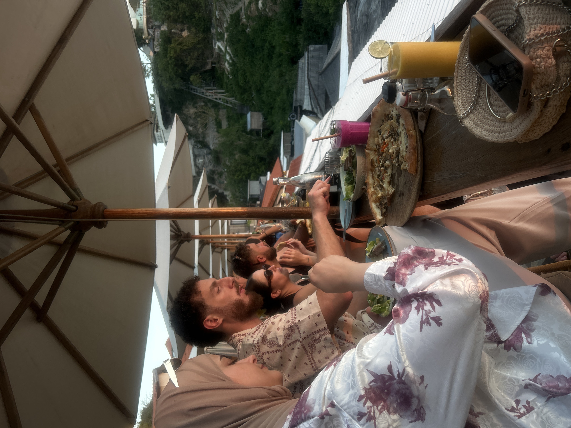 The image captures a group of people dining outdoors at a restaurant in Kuta Selatan, Indonesia, possibly at a cliffside establishment given the elevated perspective and lush green hillside in the background. The setting is characterized by large, neutral-colored umbrellas providing shade, suggesting a sunny day.
In the foreground, a woman wearing a light-colored hijab and a white robe with a floral pattern is seated. To her right, a man with curly dark hair and a beard, wearing a patterned shirt, is engaged in conversation. Further back, a woman with dark hair and sunglasses is visible, and behind her, another man is eating.
On the table in front of them are several food and drink items. A partially eaten pizza is a central feature, alongside a bowl of salad, a pink smoothie, and a yellow drink garnished with a lime slice. A glass bottle, a shaker, and a woven bag containing a smartphone are also present on the table. The overall atmosphere appears to be relaxed and social, with individuals interacting and enjoying their meal. The view beyond the table reveals the natural landscape of Kuta Selatan, with dense greenery and structures built into the hillside.