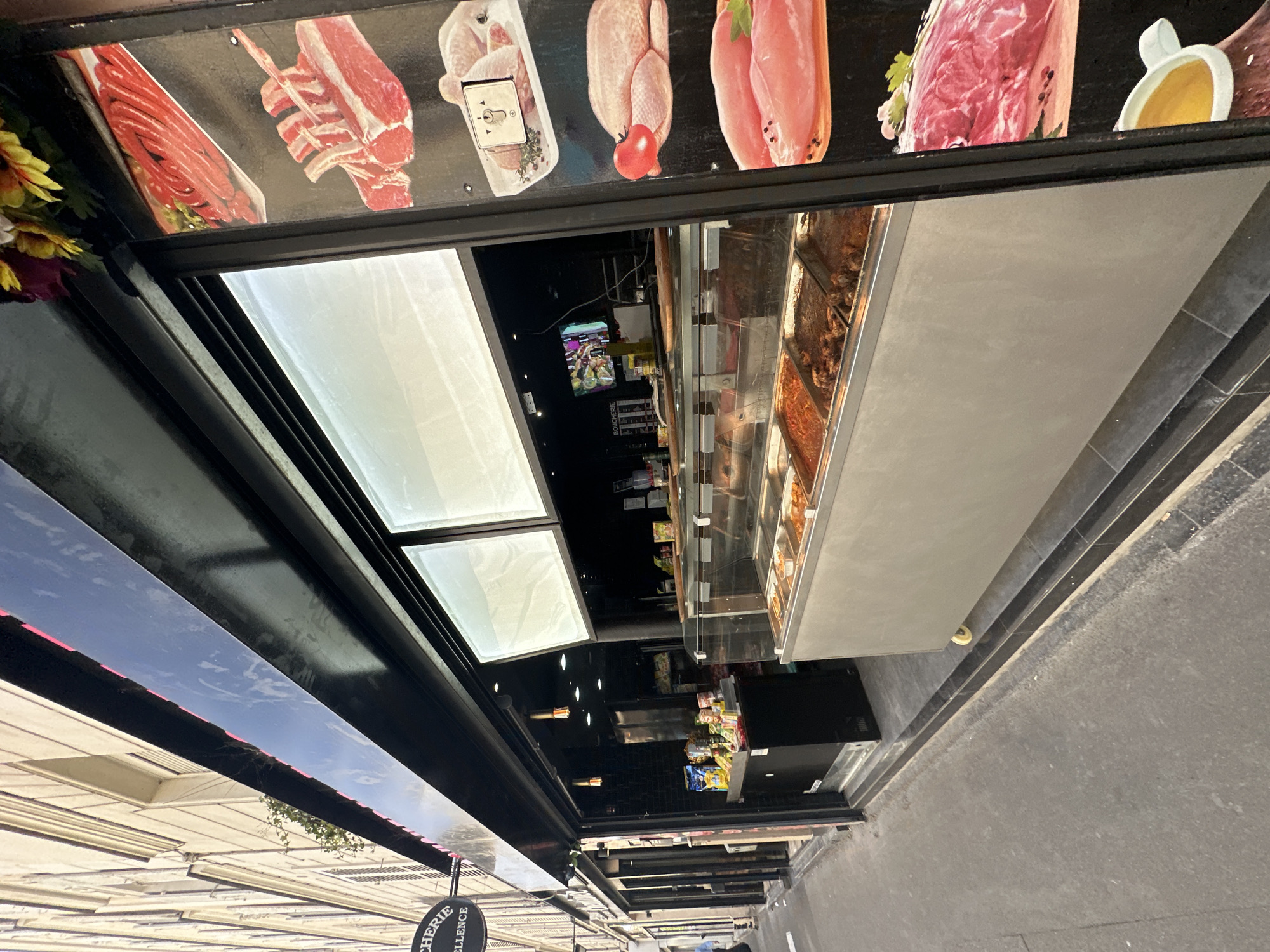A storefront, possibly a butcher shop named "Boucherie Excellence" based on a sign visible on the left side of the image, is shown in Paris, France. The facade features a black awning over a display window that reveals a refrigerated case filled with various cooked food items. To the right of the storefront, a vertical poster displays images of different cuts of raw meat and poultry. The street in front of the shop appears to be paved with asphalt, and there are buildings on the left side of the street. The lighting suggests it is daytime, with sunlight casting shadows. No people are clearly visible in the scene.