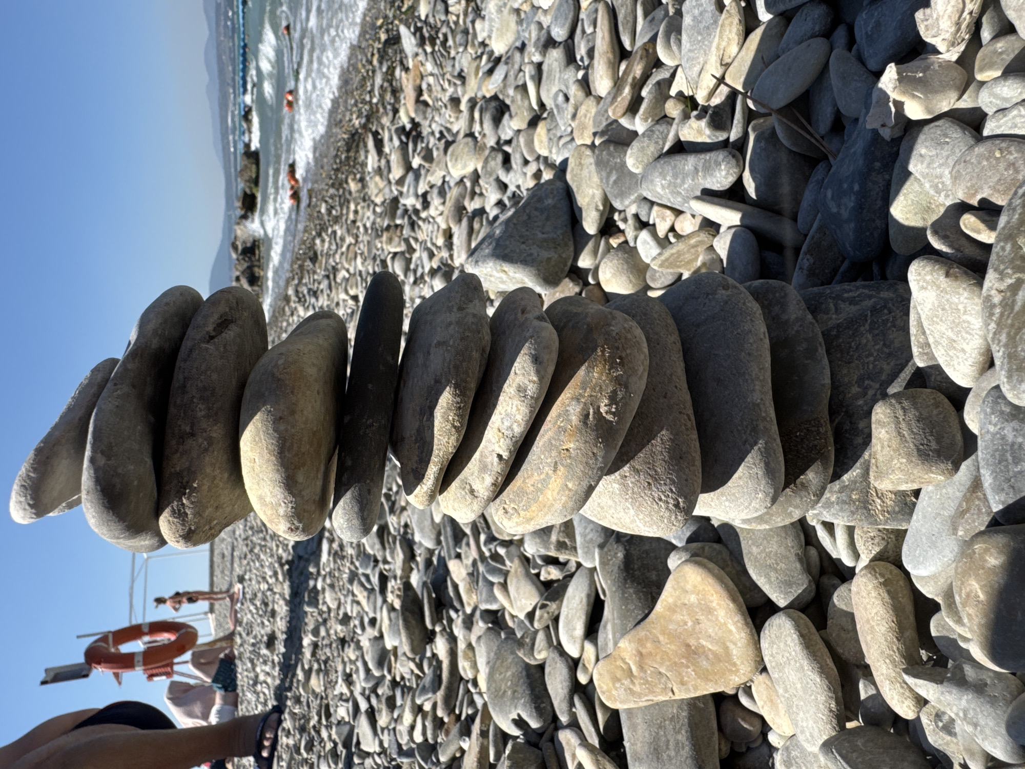 A stack of smooth, rounded stones is centrally positioned on a pebble beach in Gelendzhik, Russia, under a clear blue sky. The stones are carefully balanced on top of each other, forming a tall, narrow tower. In the background, the sea is visible with gentle waves. Several buoys are floating in the water. Further in the distance, mountains form the horizon. A few people are present on the beach; one person's legs and feet are visible in the foreground, wearing flip-flops. Another person is seated on the beach behind the stack of stones, and a third person, wearing a bikini, stands near a life preserver on a pole, suggesting a lifeguard station or a public beach facility. The overall environment is sunny and appears to be a recreational area.