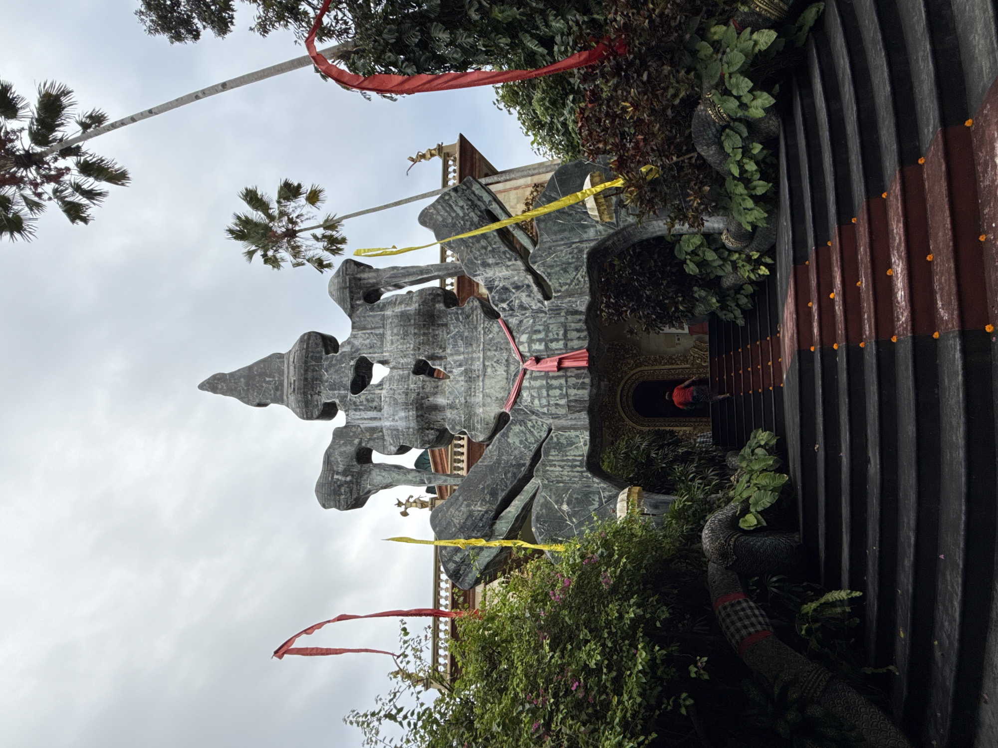 Captured in the Ubud District of Indonesia, the image presents a scene with a sky filled with clouds providing ambient lighting. A tall stone structure dominates the central space, adorned with red and yellow ribbon decorations. Lush greenery surrounds the base of this structure, featuring plants and flowers. The right side of the frame showcases a portion of a building with black and brown vertical stripes. At the structure's entrance, a figure can be seen wearing a red garment. A palm tree and other trees are visible overhead against the cloudy sky.