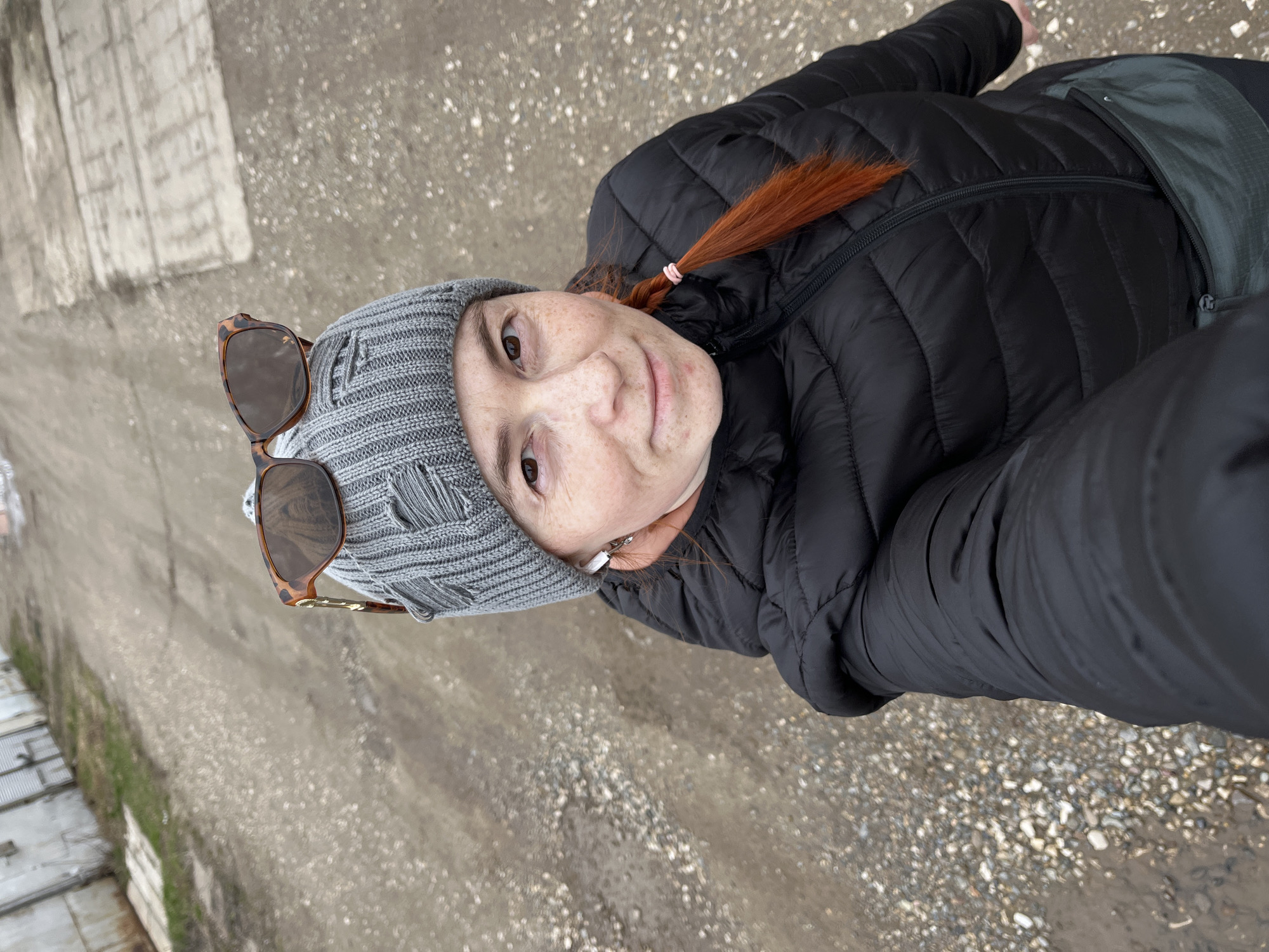 A person with fair skin, red hair, and freckles is posing for a selfie in an outdoor setting, likely the city of Chaikovsky, Russia, given the context. They are wearing a gray, knitted beanie with distressed details and sunglasses perched on top. The person is also wearing a black, puffy winter jacket. The ground beneath them is a rough, paved surface with patches of gravel and dirt. In the background, a concrete wall and some low-lying structures can be seen.