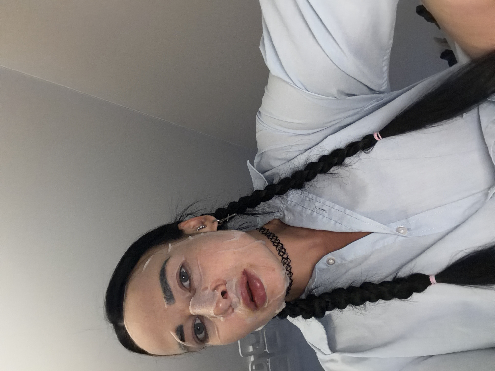 A woman with dark hair in pigtails and wearing a light blue collared shirt and a black choker is looking at the camera. She has a face mask on and appears to be in a room with grey walls.