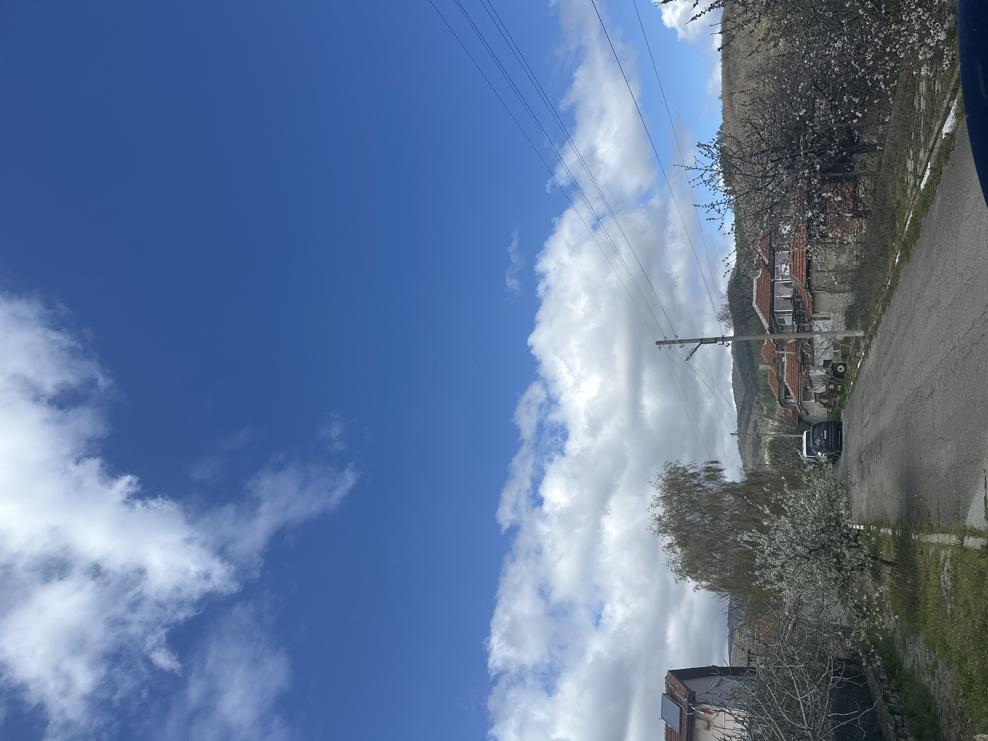 The image depicts a street scene in Gavrailovo, Bulgaria. The sky occupies the left half of the frame, showing a clear blue expanse interspersed with white cumulus clouds. Electrical power lines are visible stretching across the sky.
On the right side of the frame, a paved road runs through a residential area. Several houses with red tile roofs are visible. A black vehicle is parked along the side of the road. Trees in bloom line the roadside, with blossoms suggesting springtime. A tree in front of the house also has white blossoms. The terrain appears to be slightly hilly, with a hill in the background.