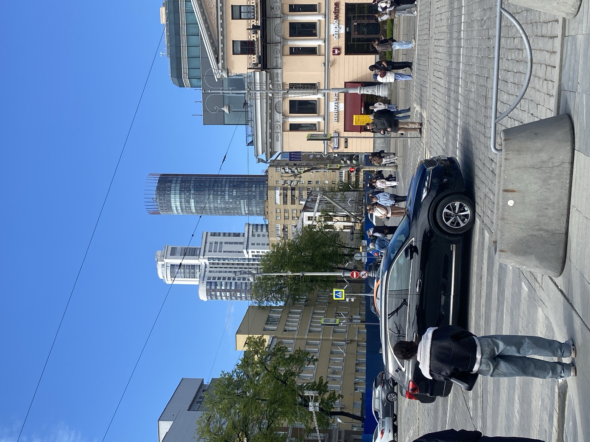 The image shows a street scene in Yekaterinburg, Russia, on a clear, sunny day. Tall buildings are visible in the background, including a modern skyscraper and other high-rise residential and commercial structures. A mix of architectural styles is evident.
People are walking on the sidewalk, some grouped together, suggesting they might be waiting or conversing. A black car is parked on the side of the road, partially blocking a pedestrian crosswalk. A person wearing a dark jacket with light-colored stripes and jeans is standing near the car.
Street signs and pedestrian signals are visible, indicating a functioning urban environment. The sidewalk and road are paved. Street furniture, such as barriers, is also visible. The presence of trees and power lines adds to the urban landscape. A store named "SimpleWine" is visible in the background.