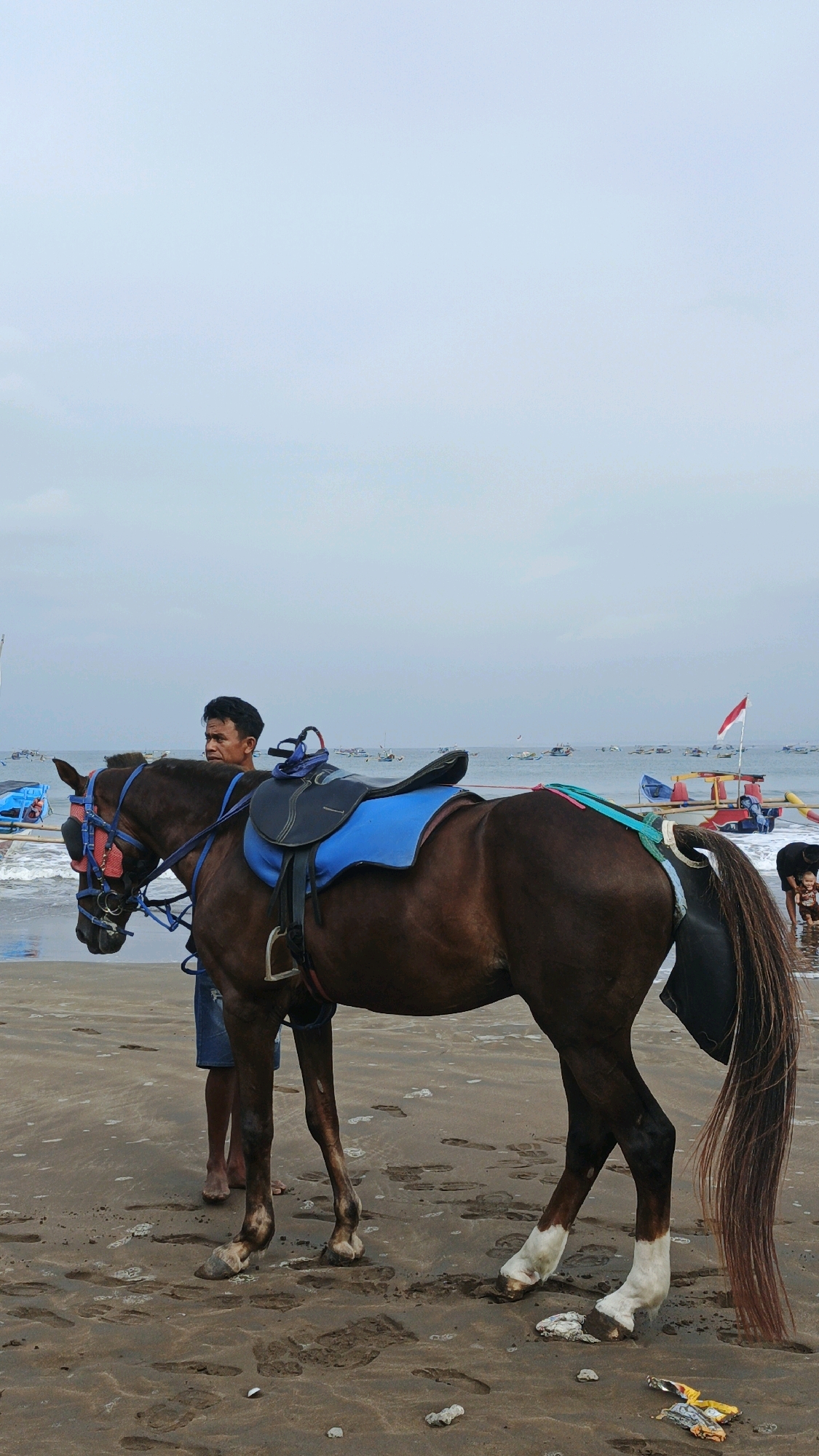 A brown horse with a blue saddle stands on a wet sandy beach in Desa Pagandaran, Indonesia, with the ocean and numerous boats in the background under an overcast sky. A man in denim shorts stands to the left of the horse, holding a blue rope attached to its bridle. The horse has white markings on its lower legs and a long, dark mane and tail. Several fishing boats are scattered across the water. A red and white Indonesian flag is visible on one of the boats. In the shallow water to the right, a person is partially visible. Footprints are imprinted in the wet sand around the horse. Debris, including wrappers and rocks, is scattered on the sand in the foreground.